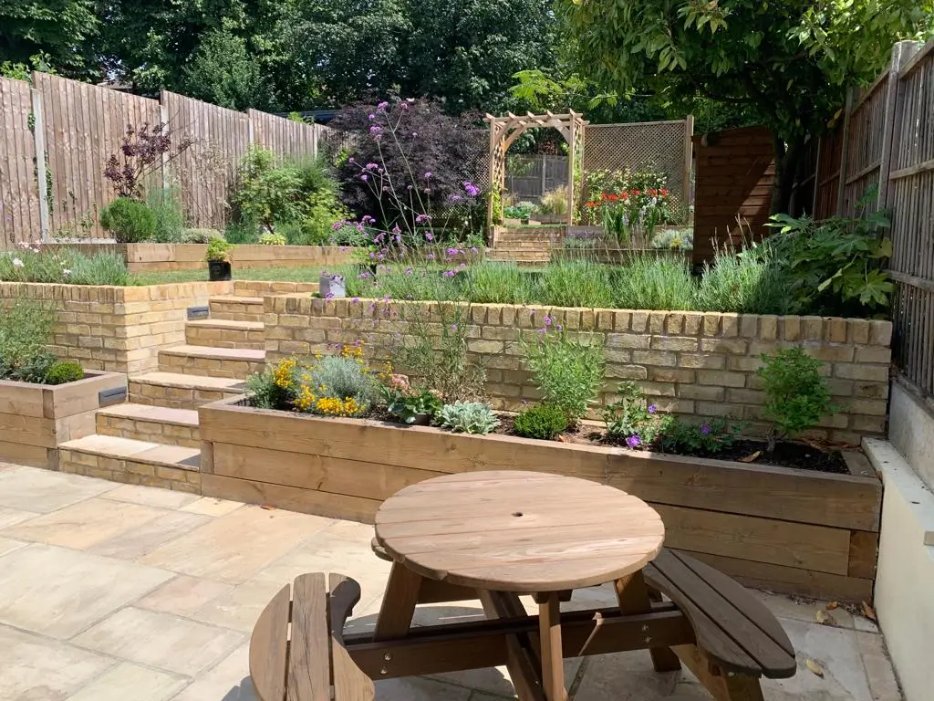 A sunny garden with raised beds, steps, and a wooden table, surrounded by various green plants and shrubs.