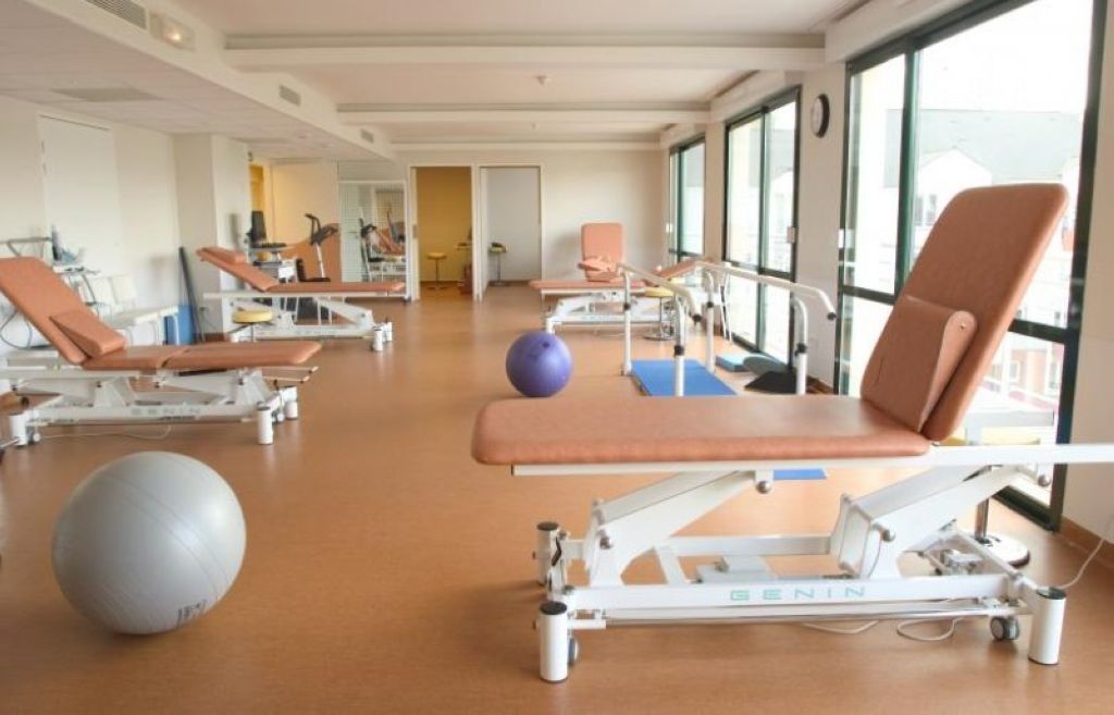 Clinical therapy room with wooden flooring, patient beds, brown leather chairs, exercise ball, and natural light from windows