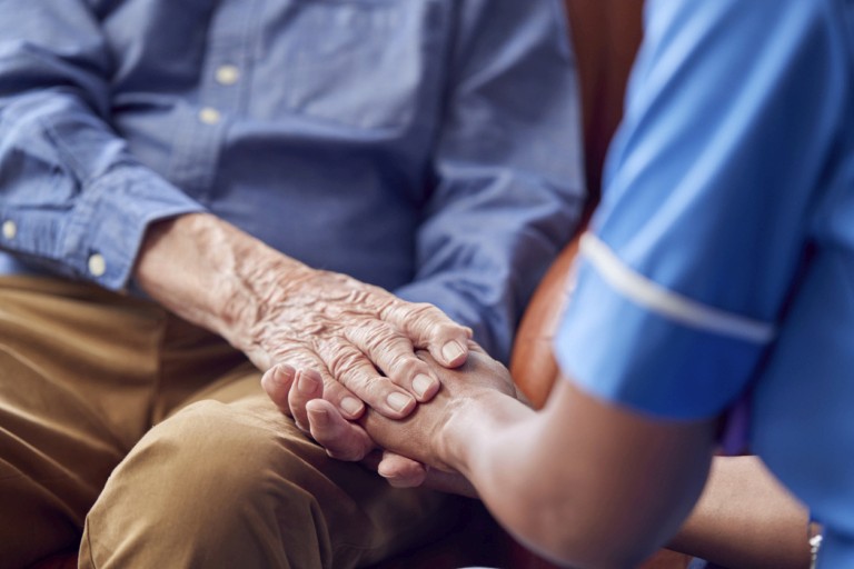 A nurse in blue uniform gently holding the hands of an elderly person, representing compassionate dementia assessment and personalised care support