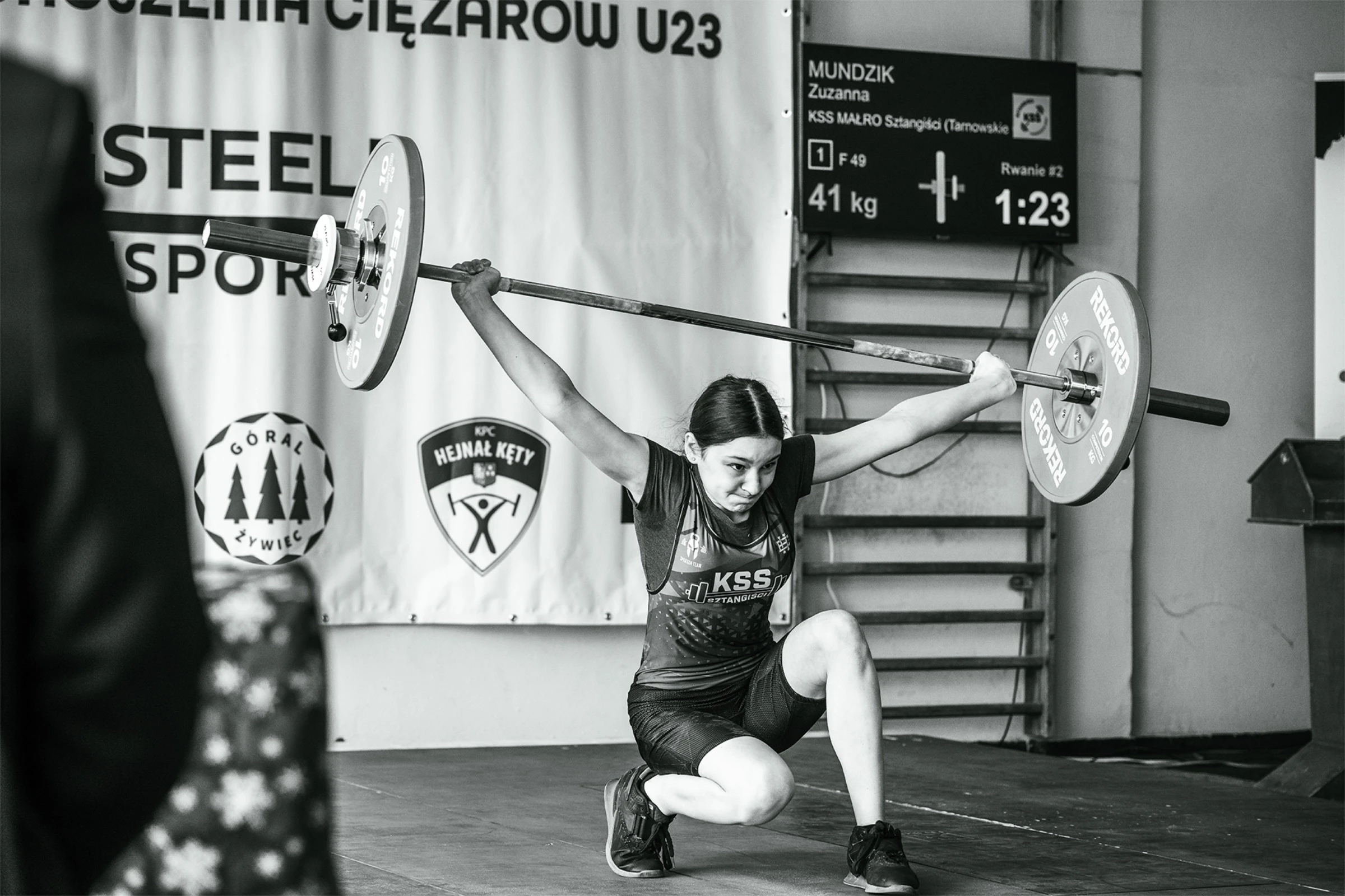 Młoda kobieta podnosi ciężar, trzymając sztangę przed ramionami. Zawody w podnoszeniu ciężarów U23.
