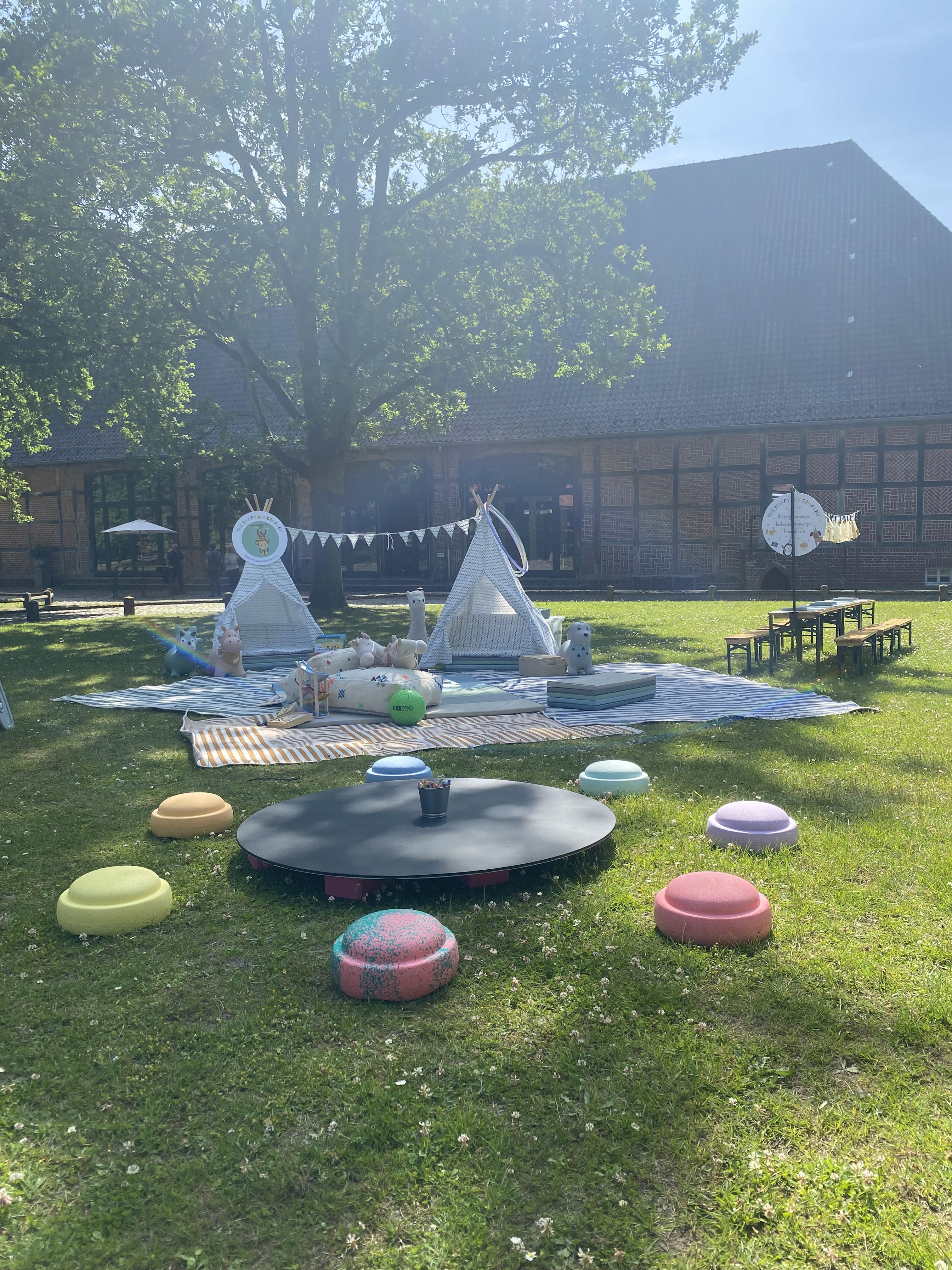 Ein Kinder-Spielbereich auf einer grünen Wiese, im Sommer in einer Hochzeitslocation. Im Vordergrund Stapelsteine und viele Spielsachen die Kindern Freude bereiten auf einen spannenden Tag.
