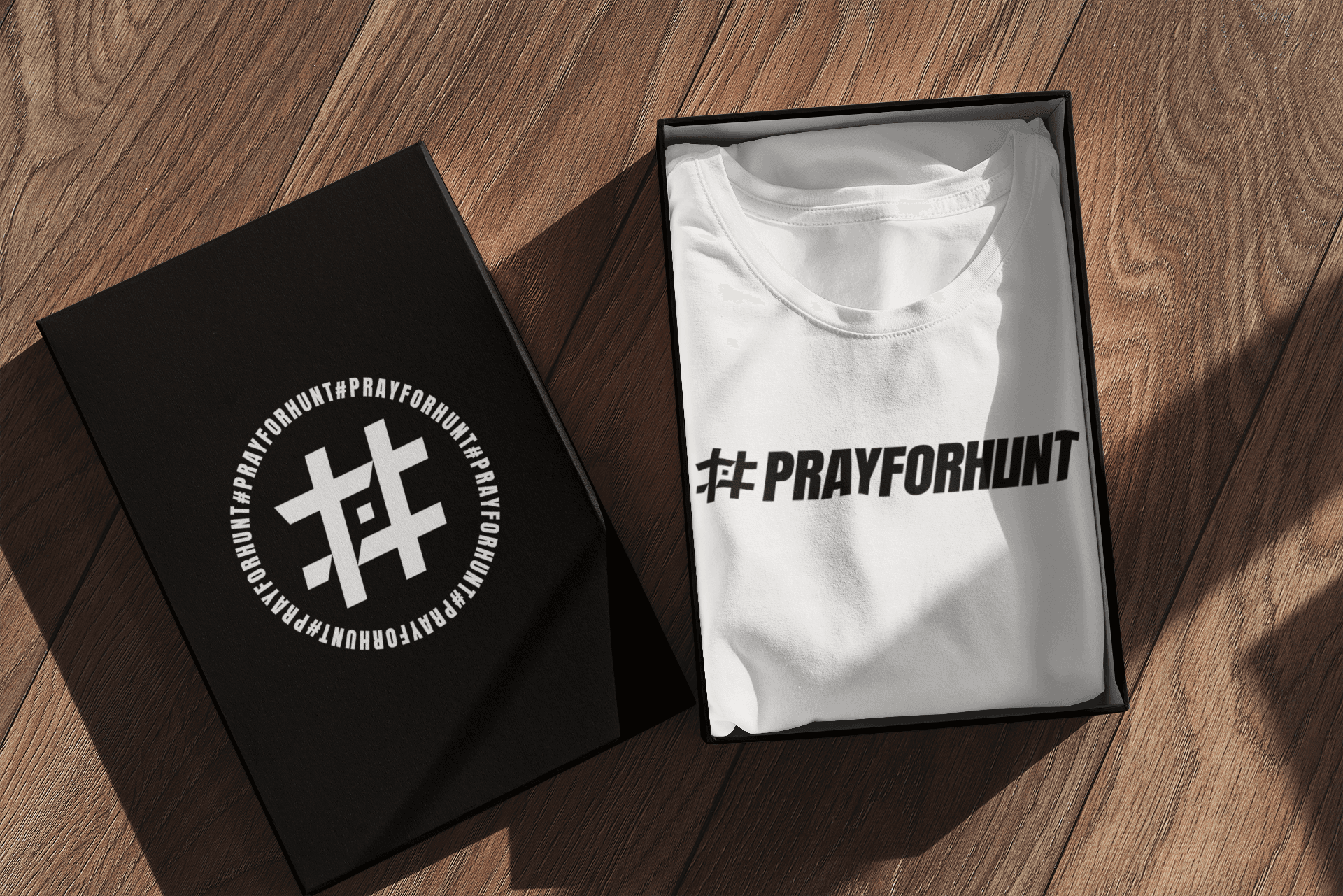 A neatly folded white T-shirt inside a black box, displayed on a wooden surface. The T-shirt features the '#PRAYFORHUNT' design printed in bold black text with a stylized hashtag symbol. Next to the open box, the box lid is shown, decorated with a circular design that includes the hashtag symbol at the center and the repeated text '#PRAYFORHUNT' around it in white. The setup is illuminated by natural light, casting soft shadows that add depth and warmth to the presentation