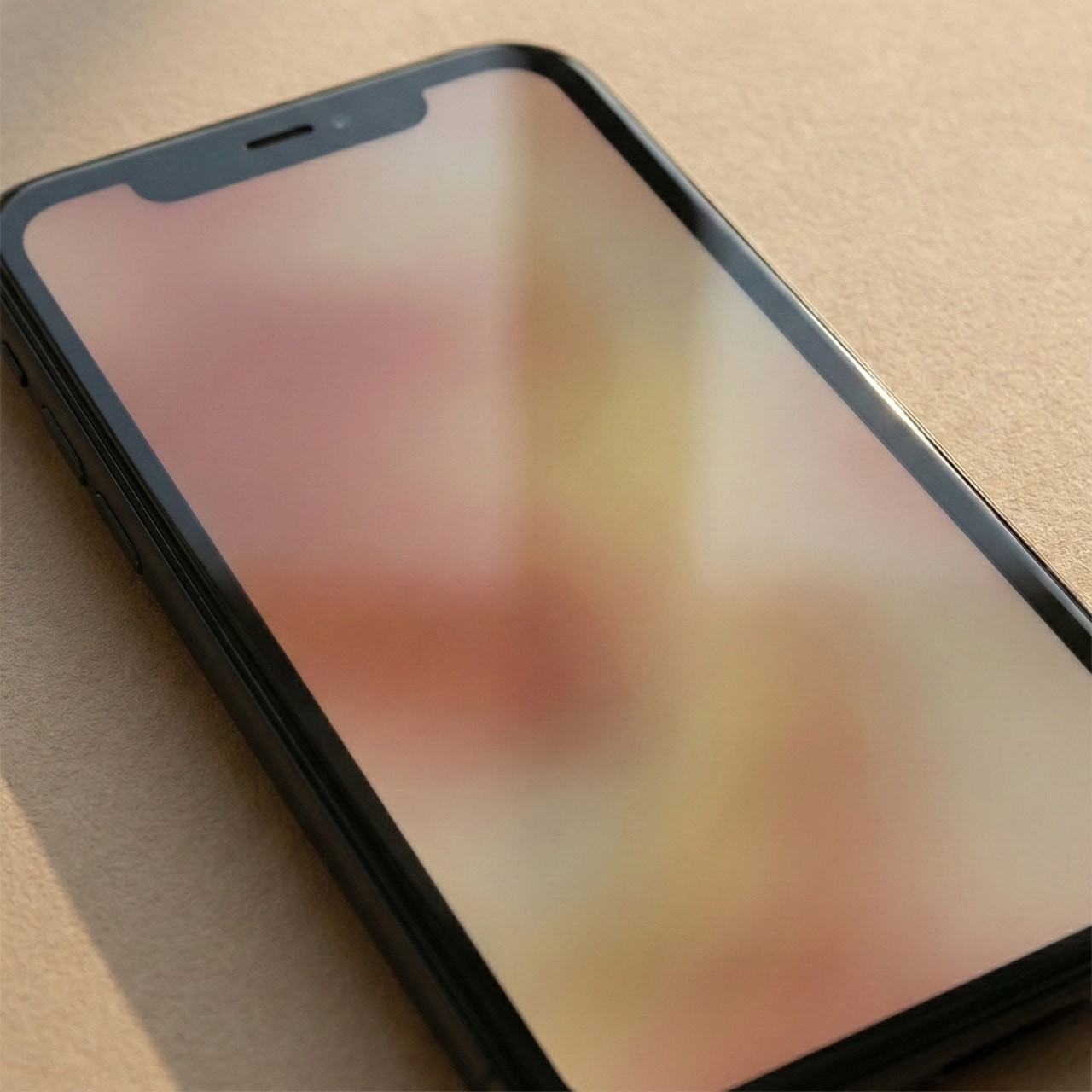 A smartphone with a dark frame lies on a light brown surface. The screen displays a blurry image with soft pink and beige tones, creating a calm, abstract feel.