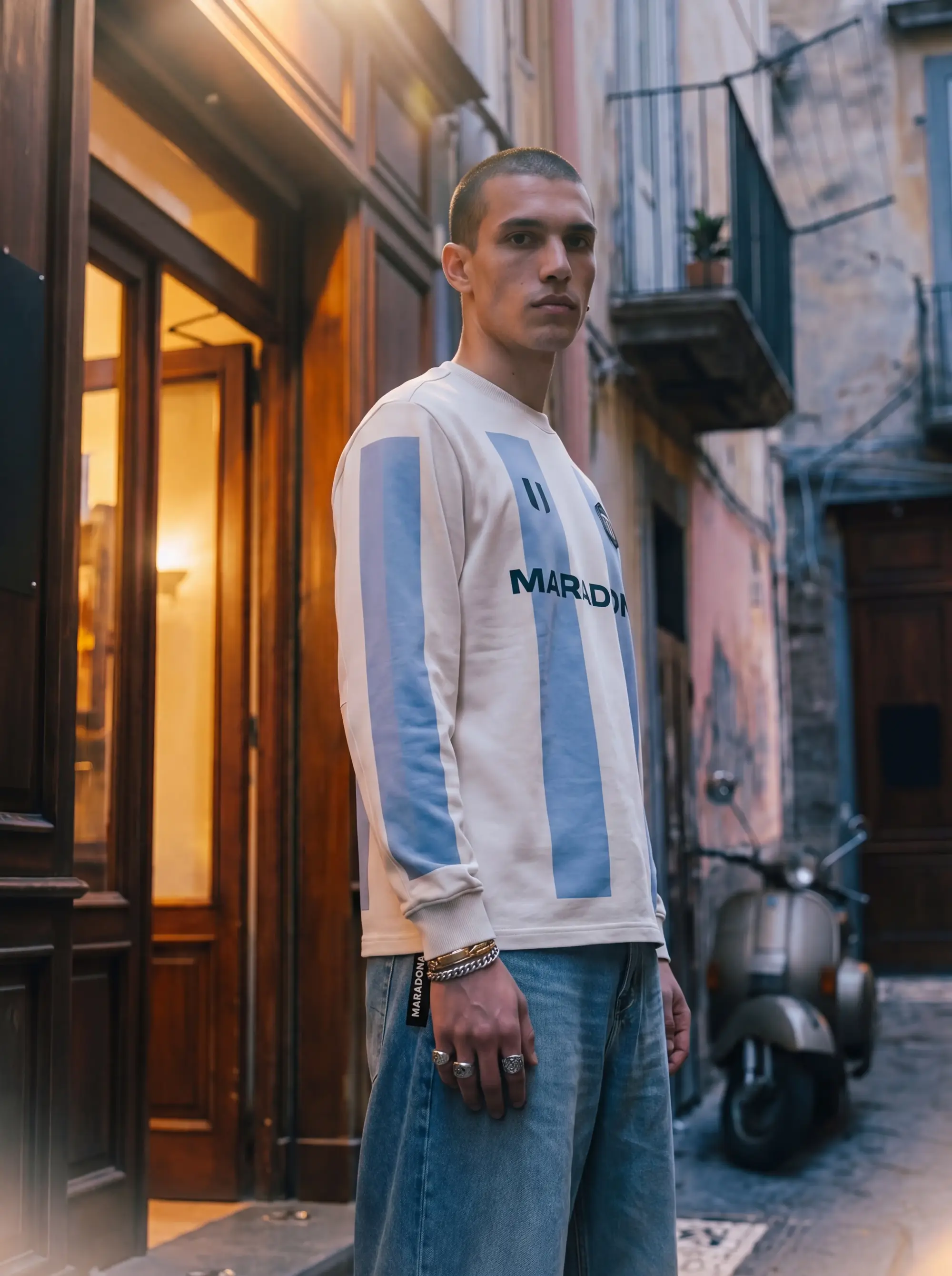 Male model wearing a cream Maradona Official long-sleeve striped shirt, standing on a cobblestone street in Napoli with a vintage scooter in the background