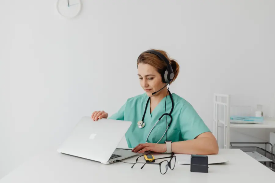 AI admin assistant for healthcare shown as a nurse in green scrubs wearing a headset while working on a laptop.