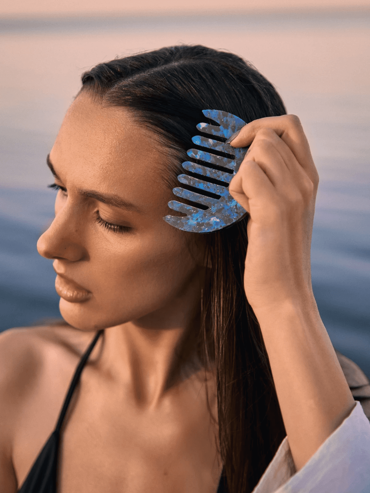 A woman with long, dark hair uses a blue, marble-patterned hair clip to style her hair, with a serene body of water and soft sunset hues in the background, while wearing a black bikini top, conveying a peaceful, beachside ambiance.