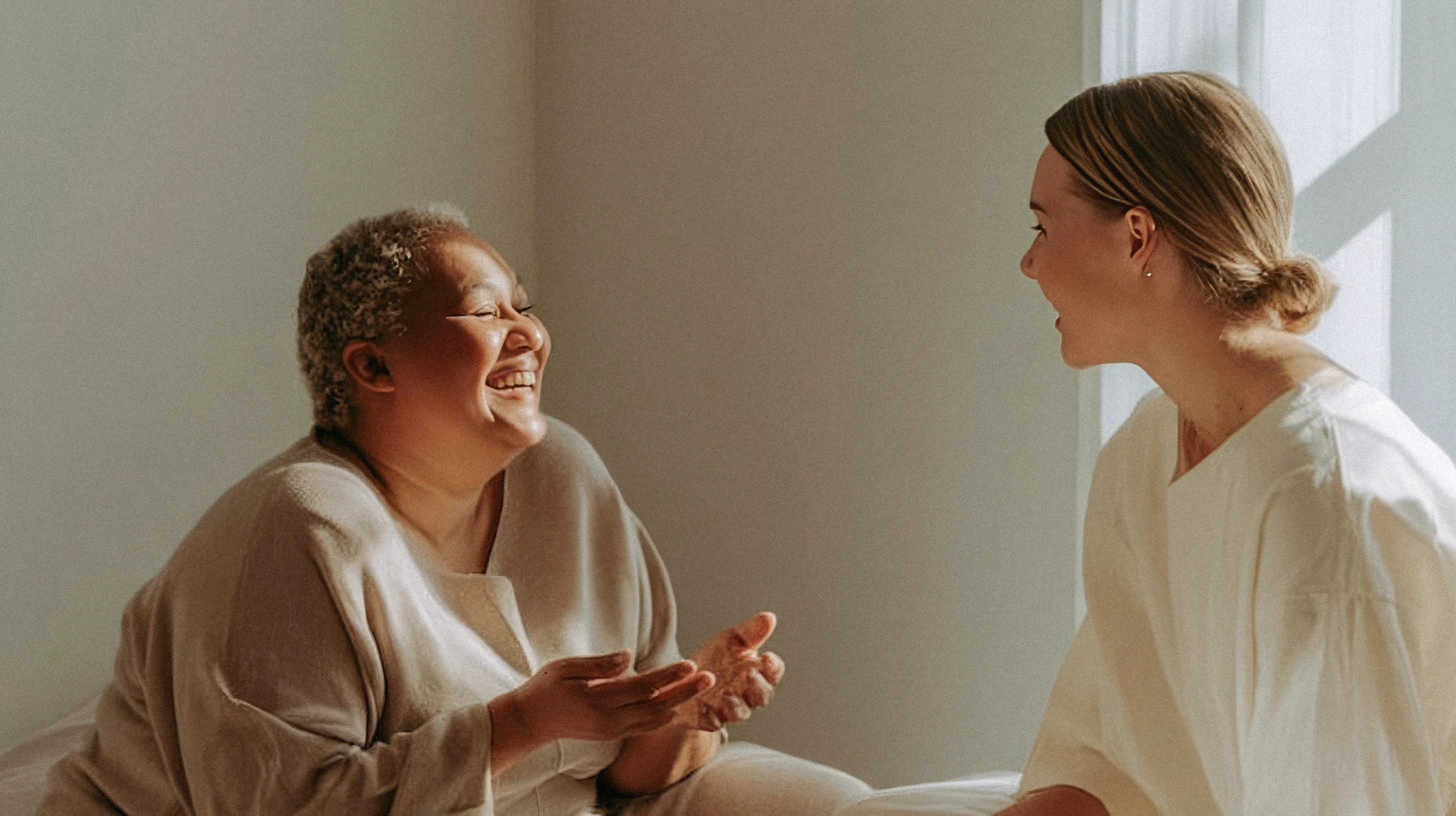 Two women sitting together, laughing warmly during a relaxed conversation bathed in soft window light.