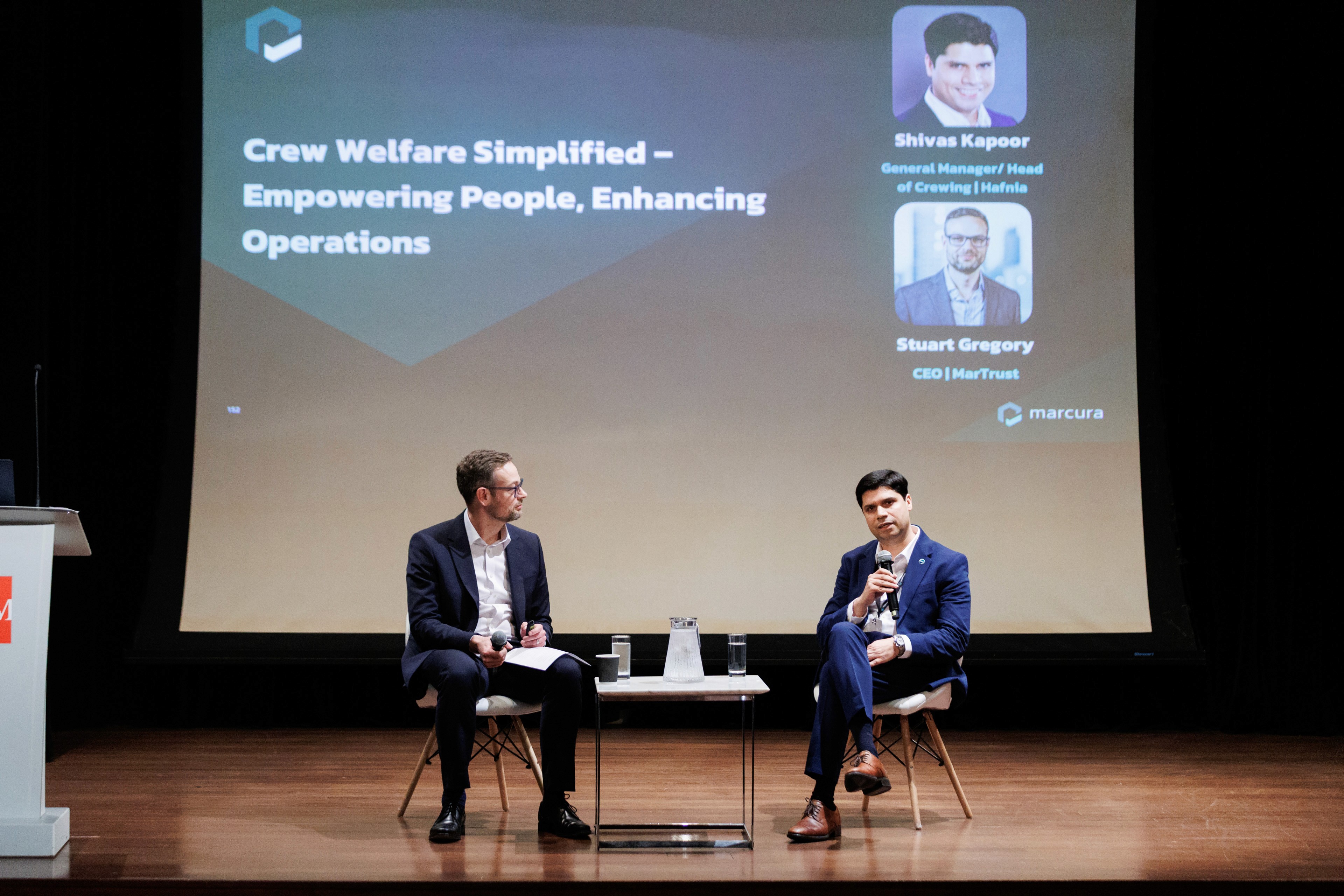 Two speakers seated on stage in a panel discussion, with a Marcura slide titled 'Crew Welfare Simplified' behind them