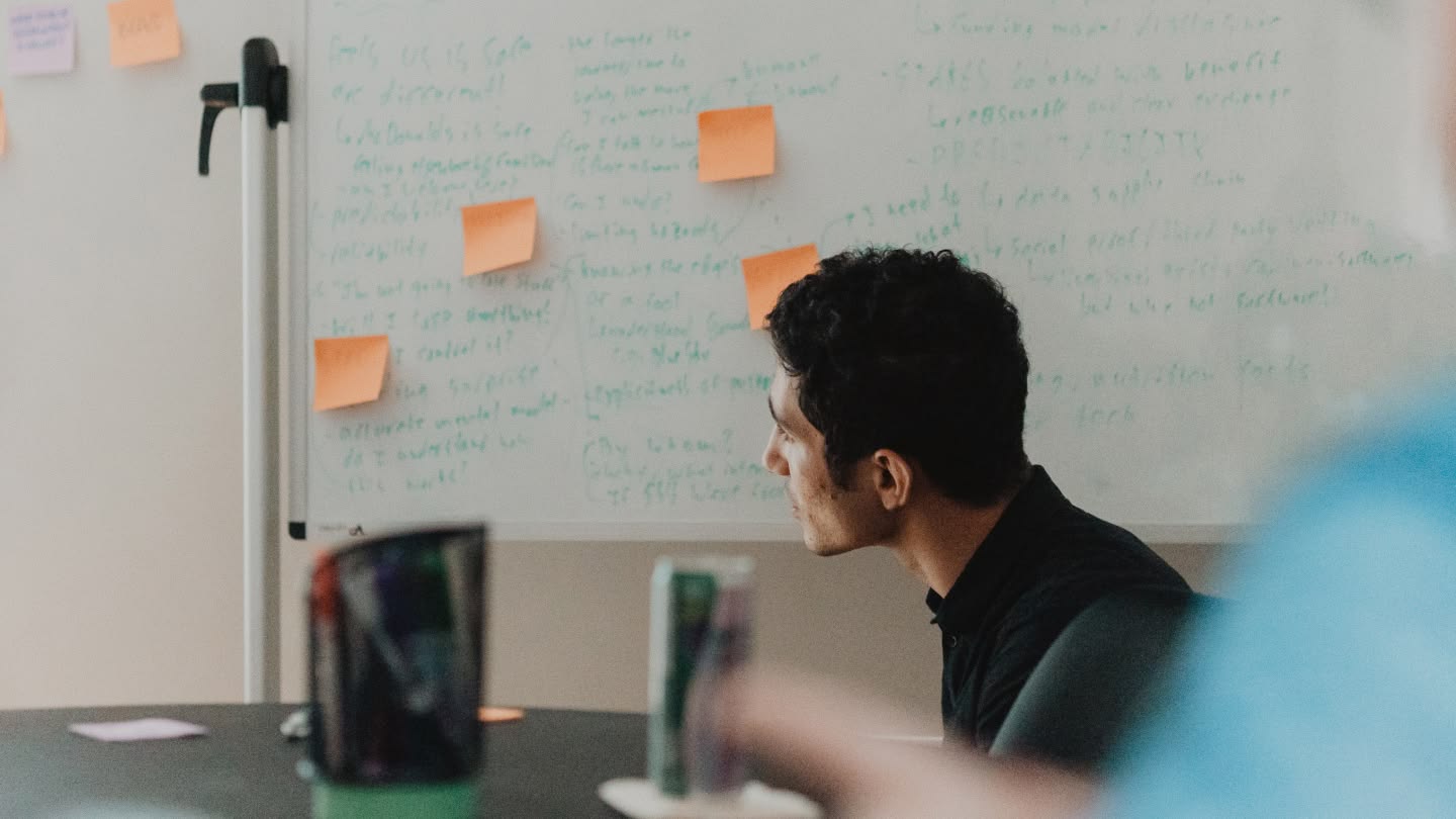 Man attentively looking at a whiteboard filled with handwritten notes and sticky notes in an office setting. The scene conveys focus and concentration.