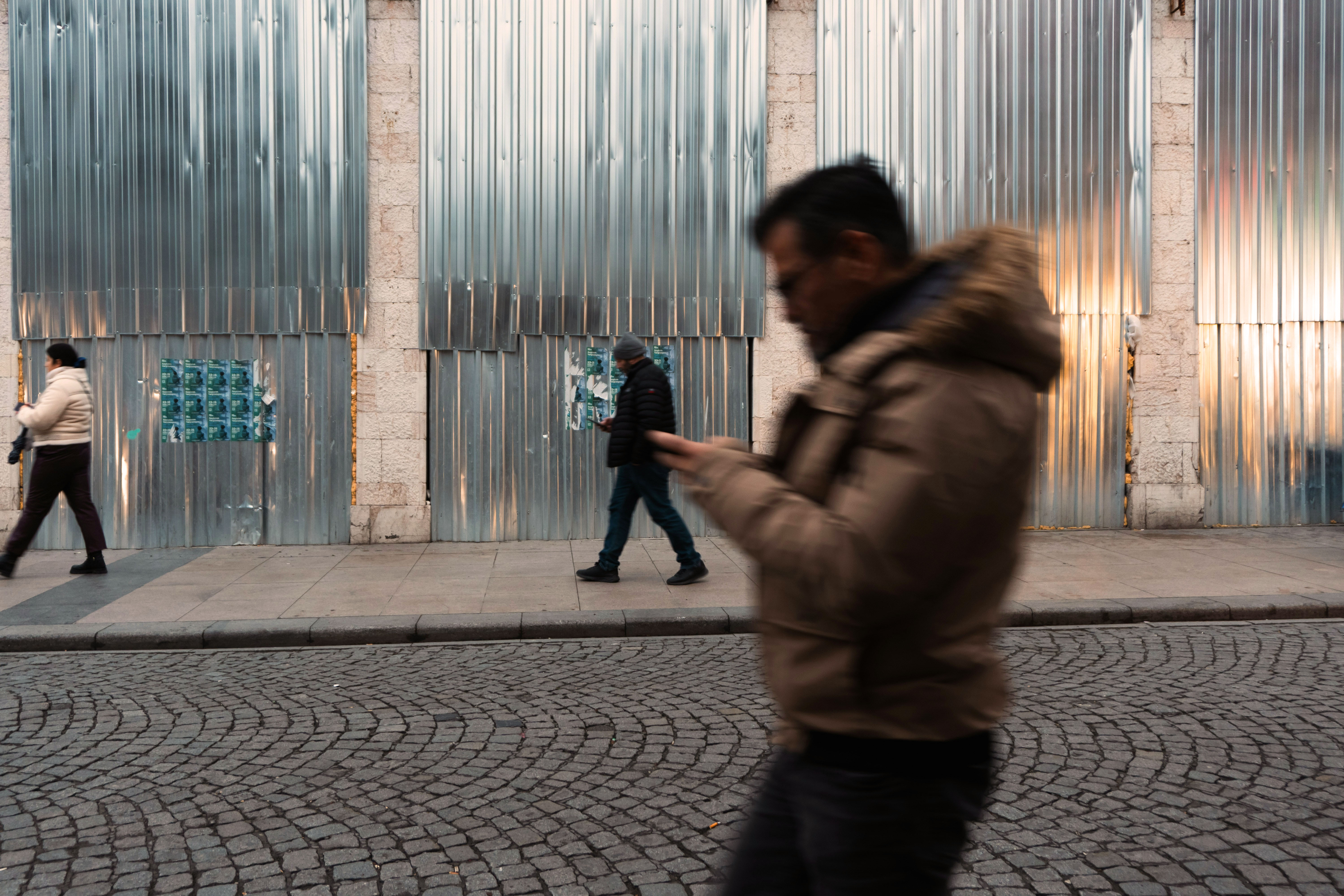 A person in a brown jacket uses a smartphone, with blurred figures walking in the background on a city street.