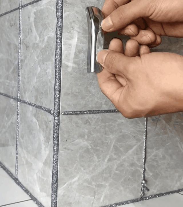 A hand holding a tool to smooth out silver glitter grout between tiles on a grey stone surface. The grout lines are being refined for a clean and polished look.