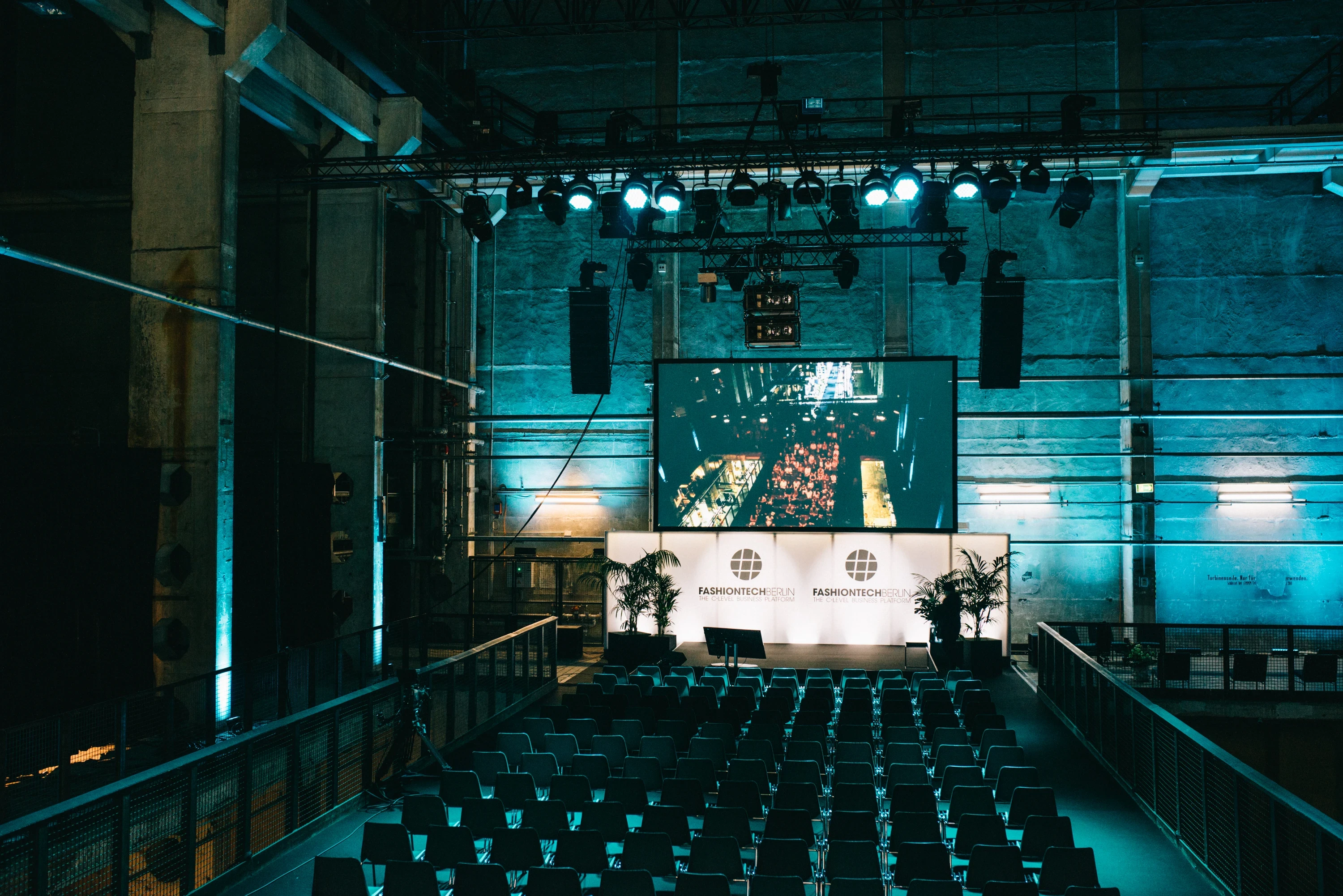 Empty conference stage at FASHIONTECH Berlin with rows of dark chairs, large screen and teal-lit concrete walls under stage trusses.