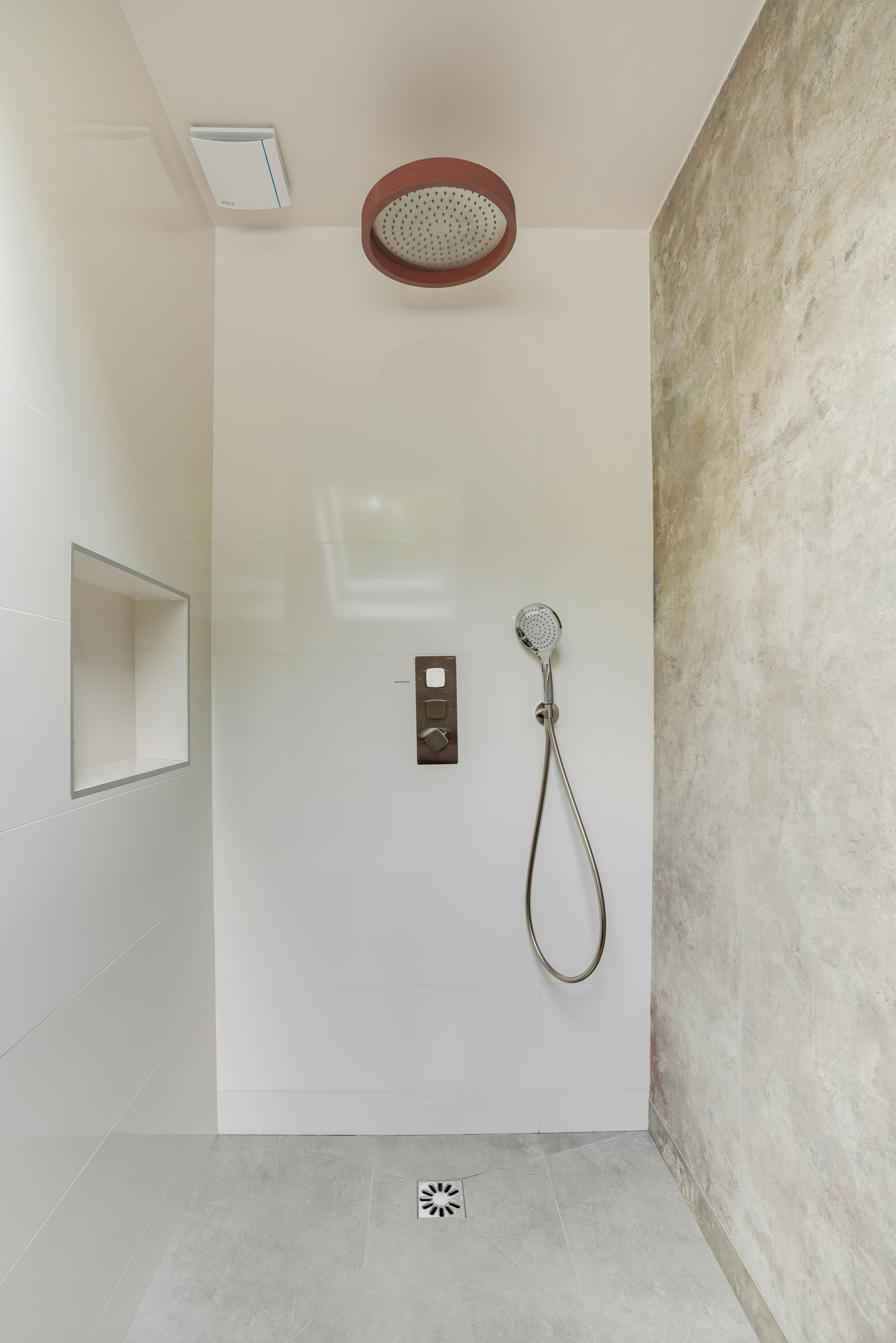 Rénovation douche minimaliste 77 avec grand pommeau plafond, murs pierre texturée et carrelage gris clair, architecte d'intérieur clé en main.
