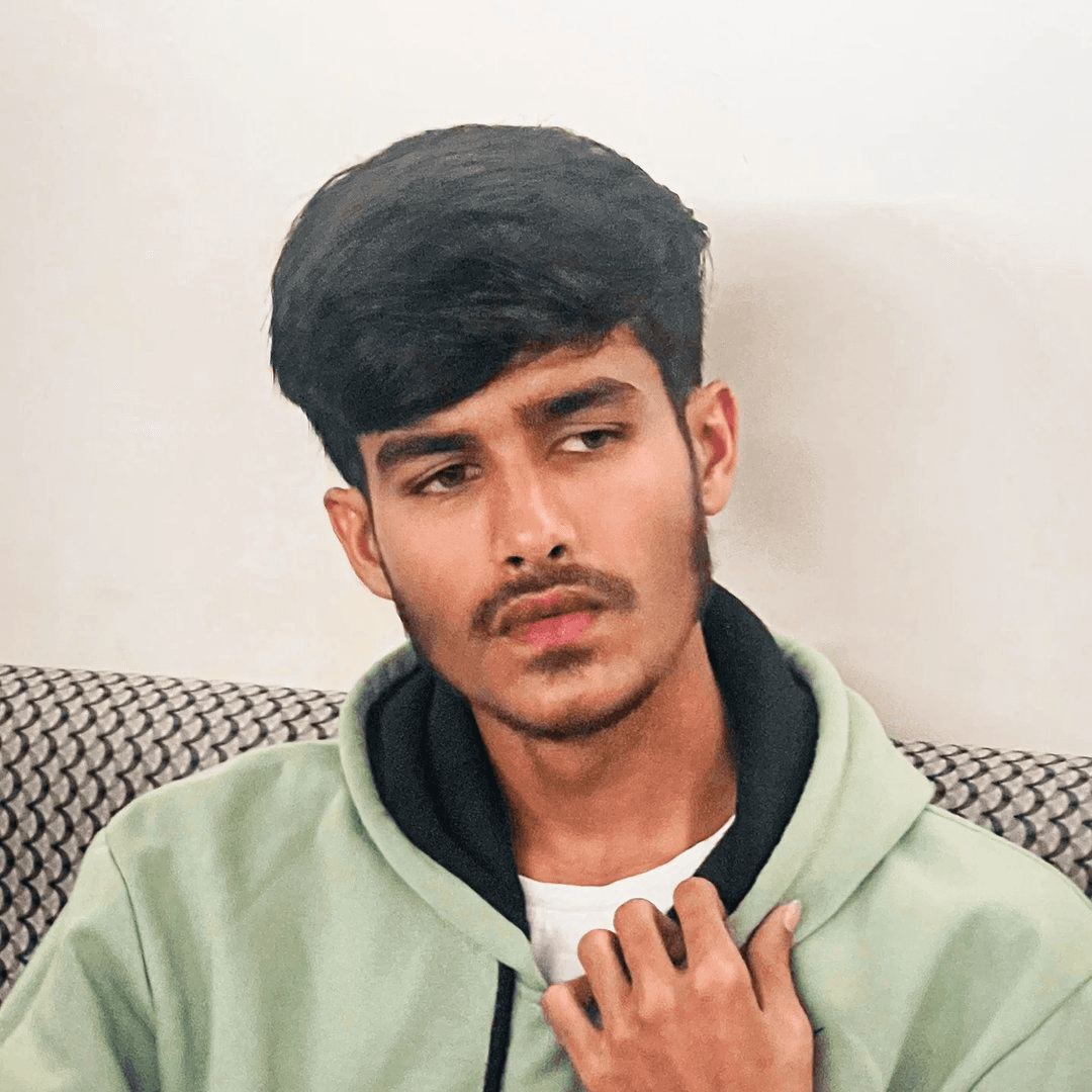 Young man with wavy hair wears a dark overcoat, rust-colored sweater, white shirt, and patterned tie, exuding a poised and sophisticated demeanor.