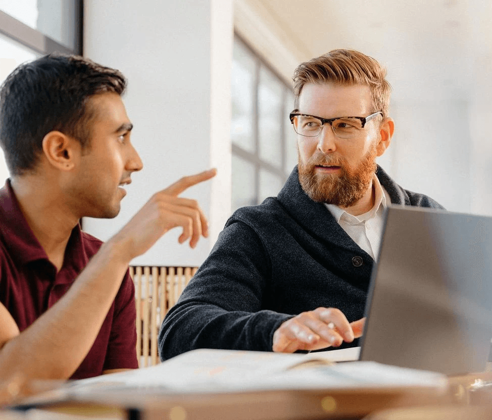 Two men collaborate in a bright office; one points and talks as a bearded colleague in glasses works on a laptop.