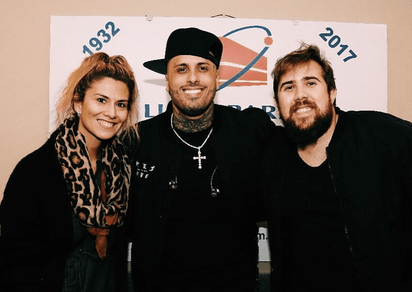 Mujer con bufanda animal print, hombre con gorra negra y collar de cruz, y hombre barbudo posan sonrientes ante un banner “1932 – 2017”.