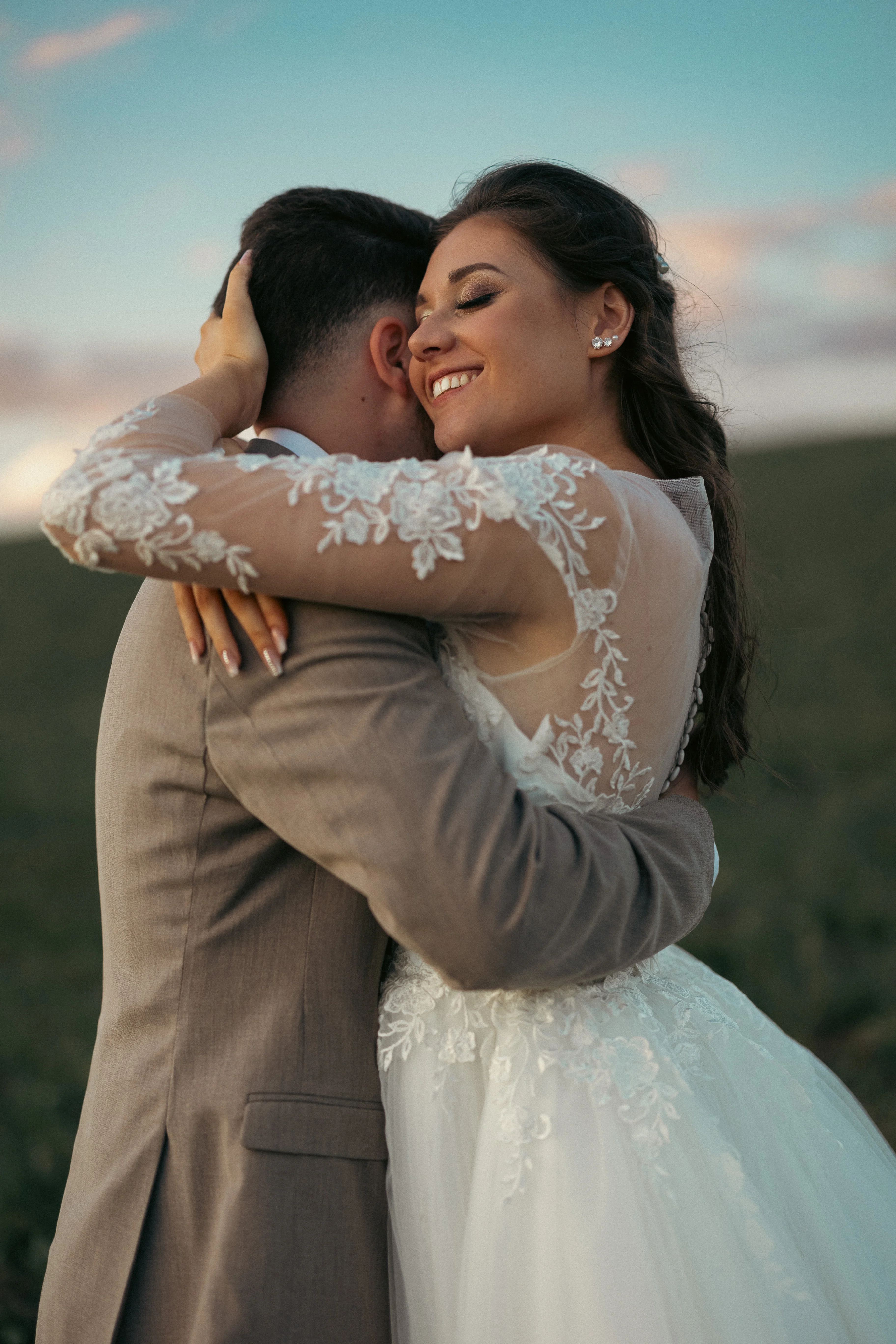 Menyasszony és vőlegény szorosan ölelik egymást, mezőn, meghitt pillanatban, természetes háttérrel.