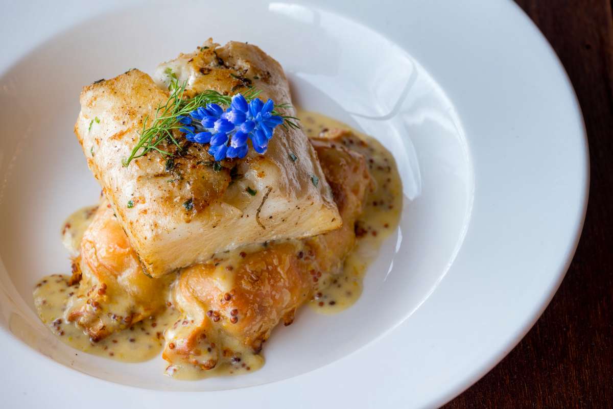 A piece of roasted fish topped with herbs and a small blue flower sits in a creamy sauce on a white plate.