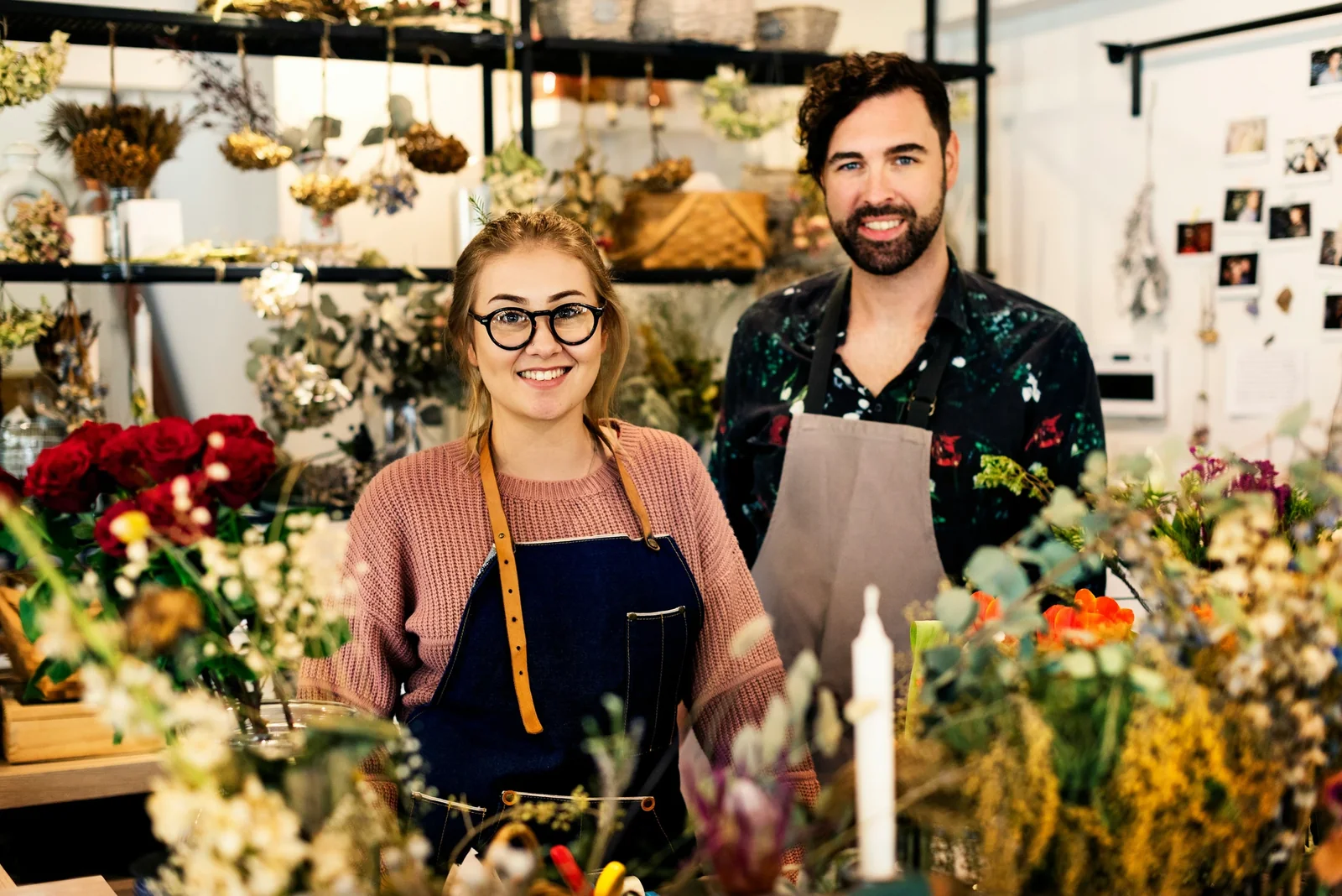 Two florists smiling in their flower shop surrounded by colorful blooms and hanging dried flowers.