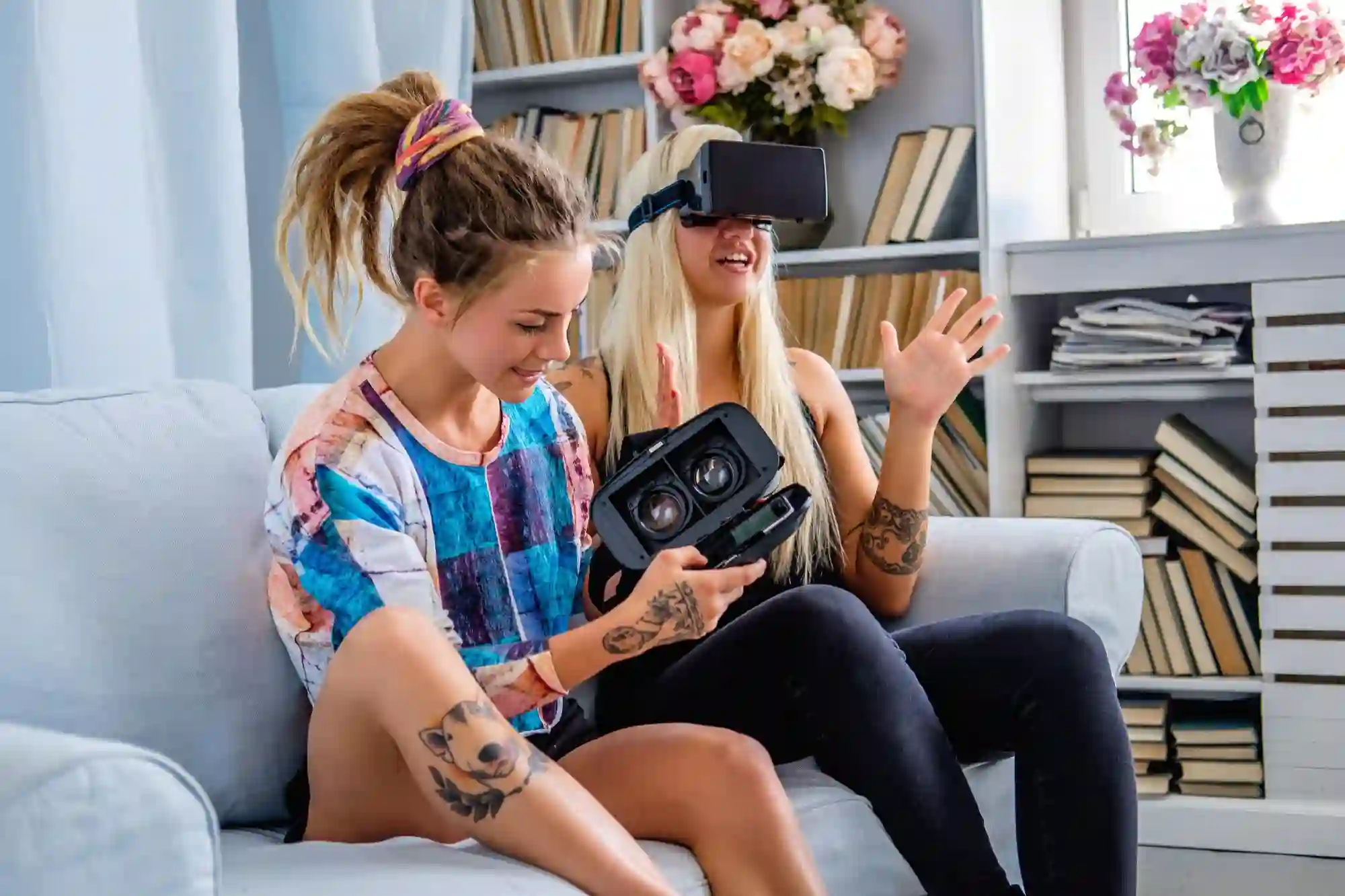 Two young women are sitting on a couch in a living room, one is wearing a VR headset and gesturing with her hands, and the other is holding and looking at a VR headset.