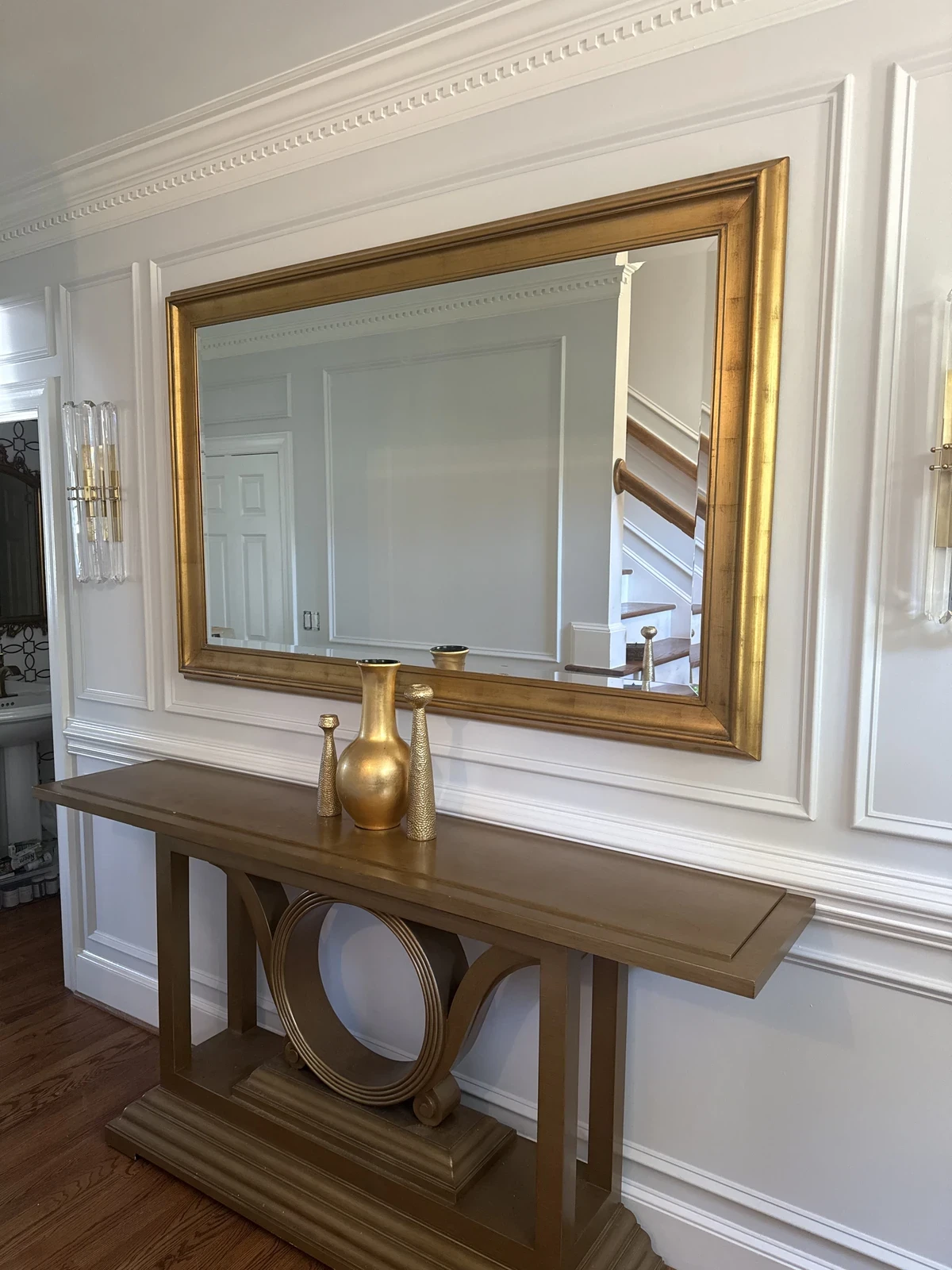 Large gold-framed mirror installed on a white paneled wall above a gold console.