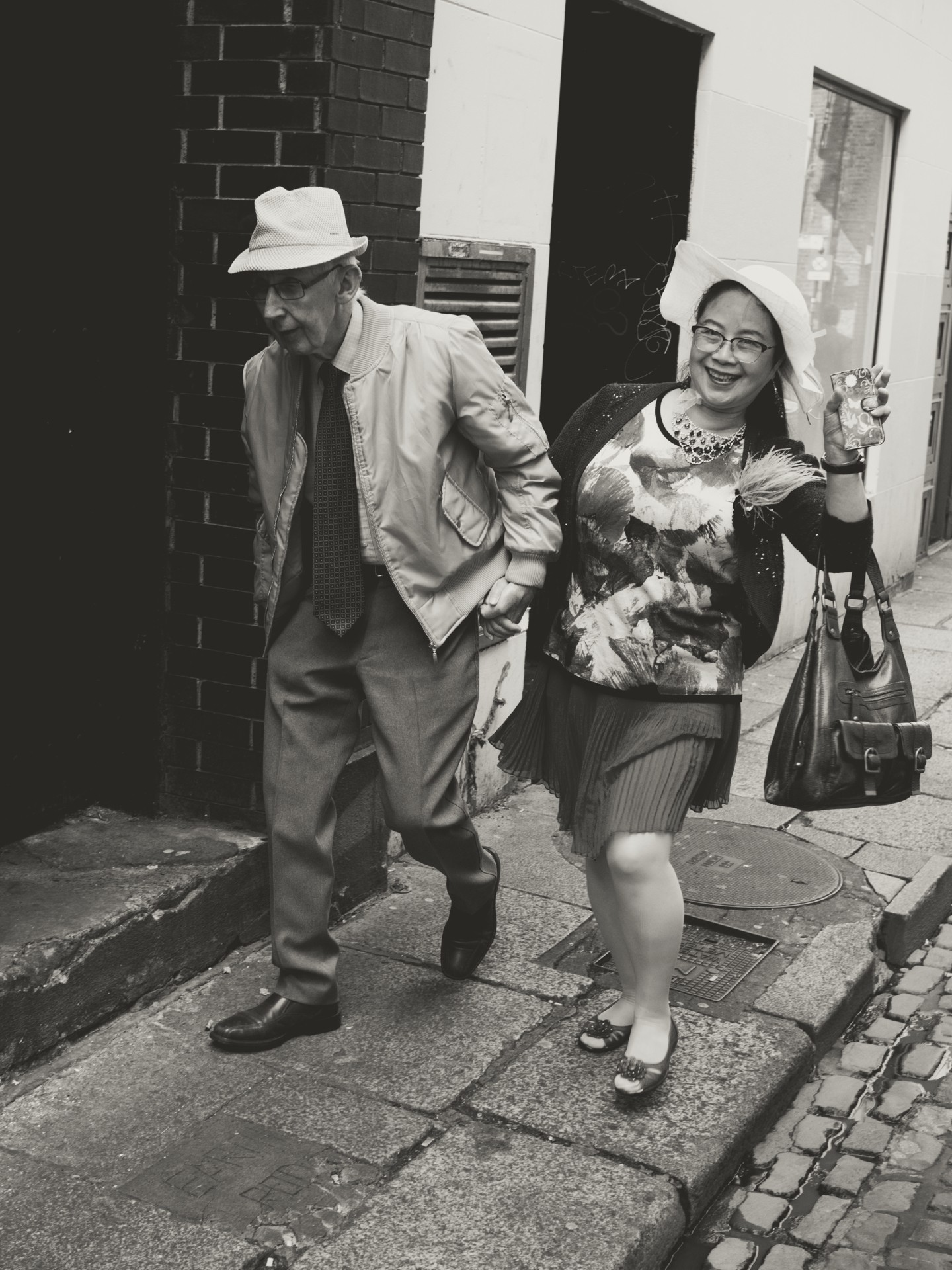 Elderly couple walking on sidewalk, man in suit and hat, woman holding purse and phone.