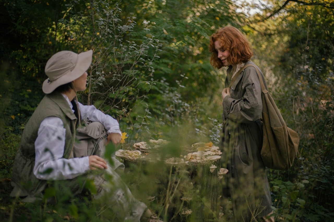 Two young girls exploring a heavily treed, forest-like garden, with sunlight filtering through the canopy.