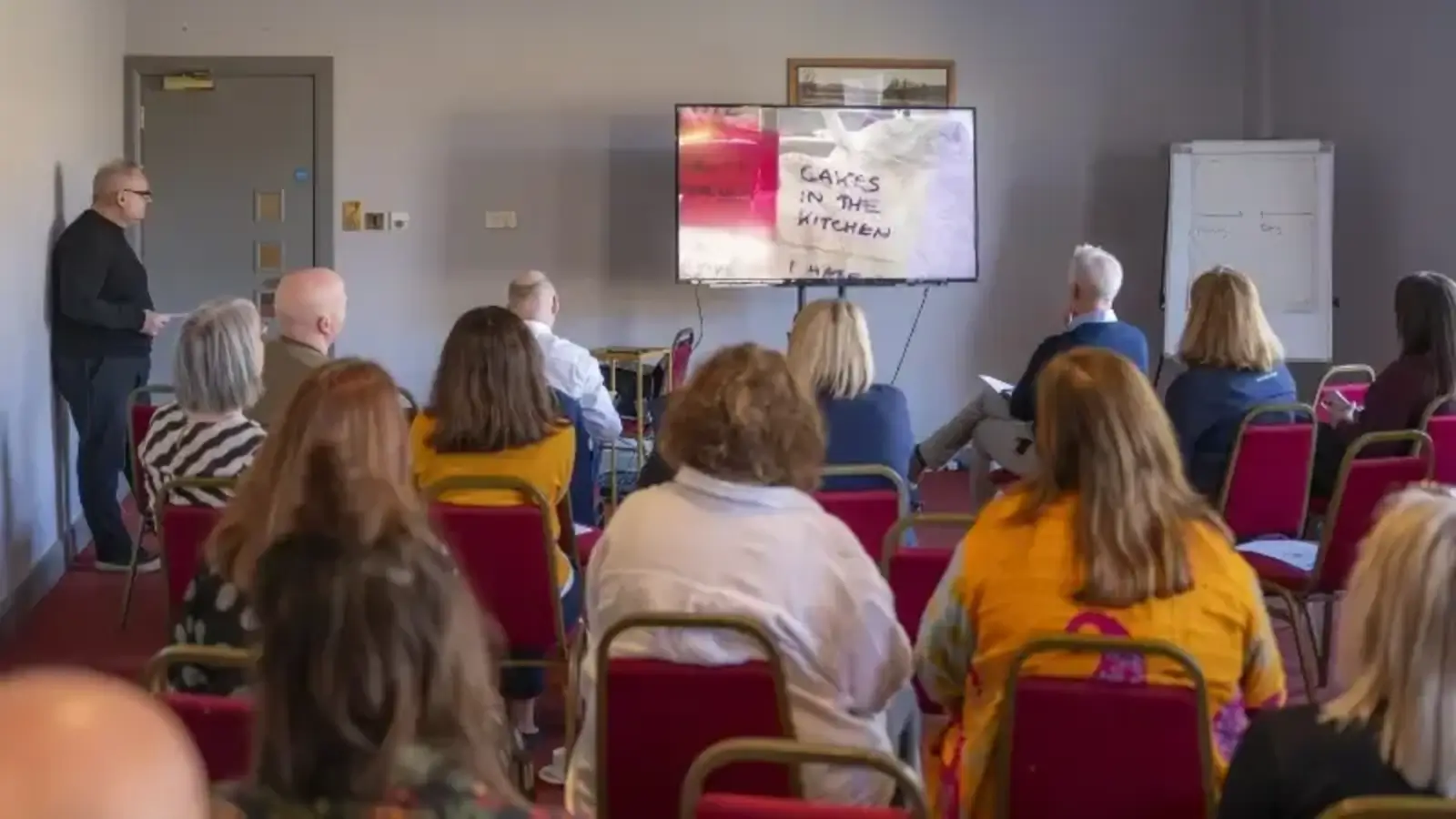 A group of people sits in red chairs facing a presentation screen in a conference room. A speaker stands to the side. The screen displays "Cakes in the Kitchen." The atmosphere is focused and attentive.