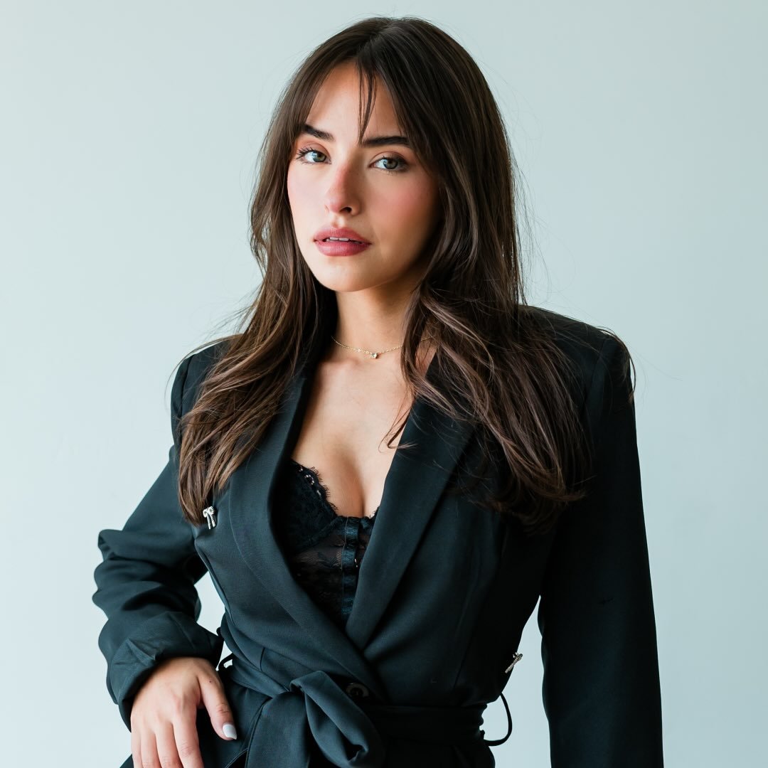 Smiling man with windswept red hair outdoors, wearing a black jacket and green backpack. Bright sunlight and a blurred natural background convey joy and adventure.