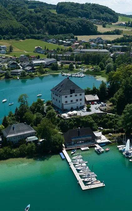 Luftaufnahme vom Schloss Mattsee, umgeben von klarem, türkisfarbenem Wasser