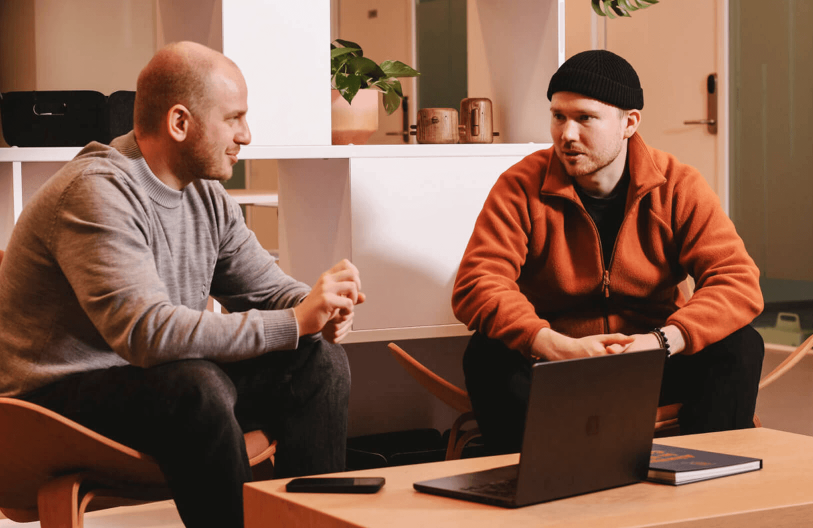 having a focused conversation in a modern office space, one wearing a grey sweater, the other an orange fleece jacket, with a laptop on the table between them.