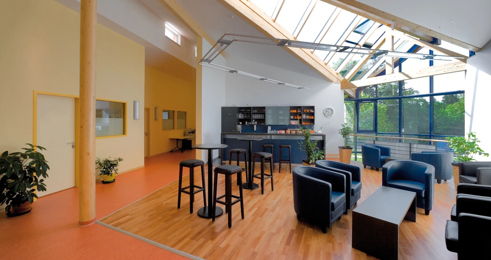 Moderner Aufenthaltsbereich mit Dachfenstern, einer Bar, Sitzgelegenheiten und Holzböden, durchzogen von einer orangefarbenen Fußbodenzone.