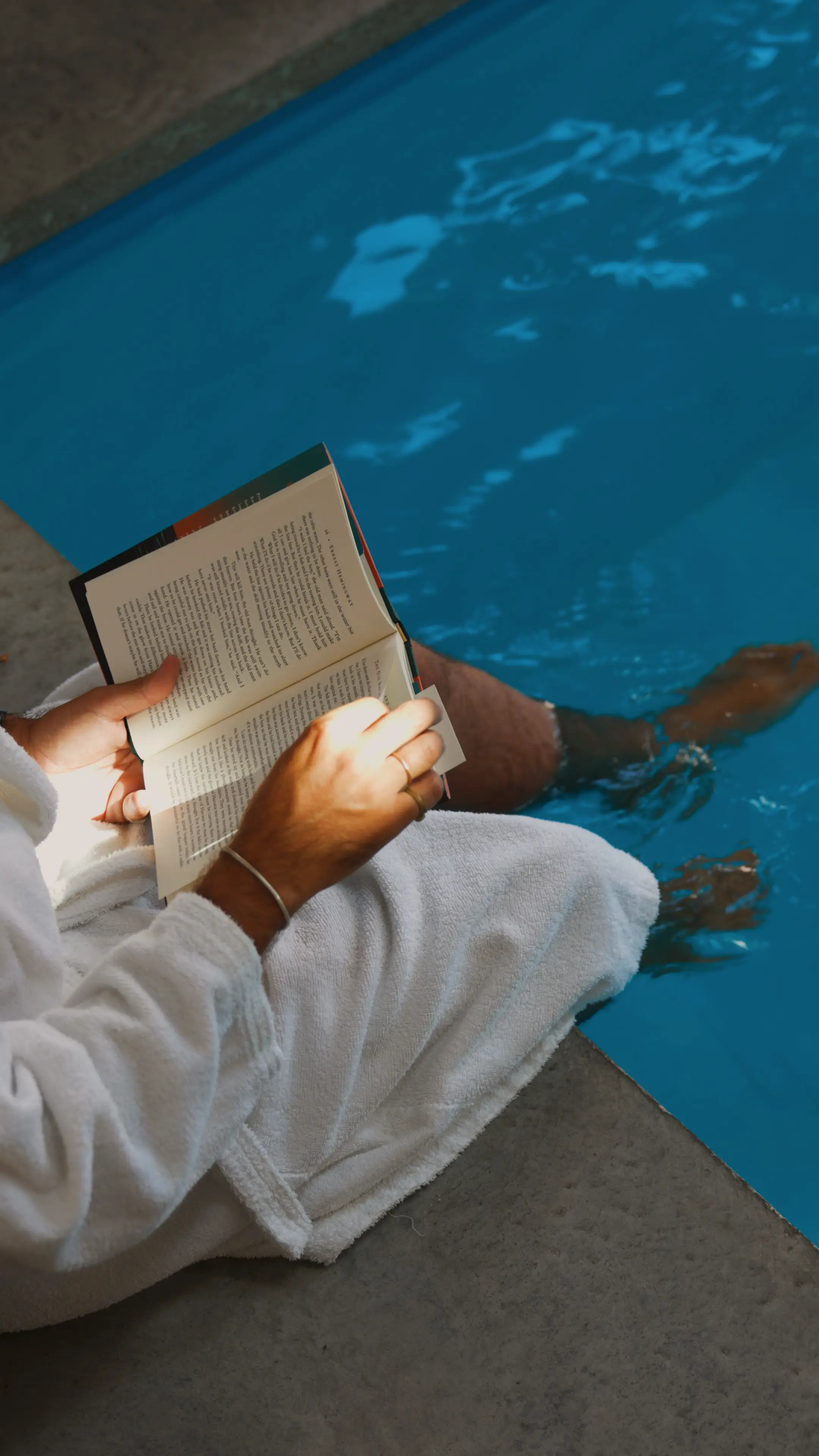 Persona en albornoz leyendo un libro con los pies metidos en la piscina