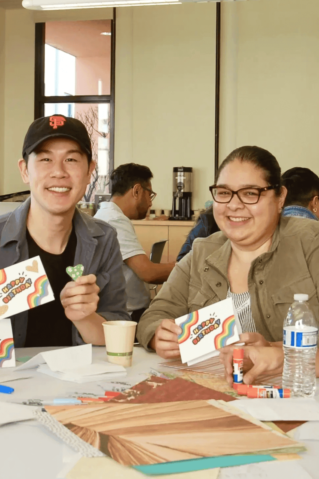 Dos personas sonríen mientras están sentadas en una mesa cubierta con materiales para manualidades, incluyendo papel de colores, tijeras, barras de pegamento y tarjetas con diseños de arcoíris, en una habitación luminosa con varias otras personas al fondo.