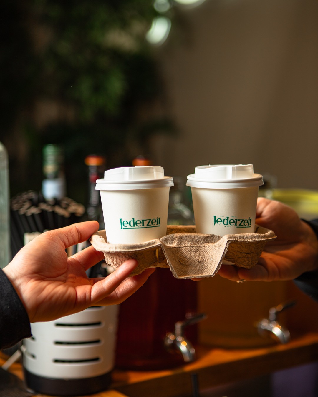 Hände halten einen Pappträger mit zwei weißen Kaffeebechern zum Mitnehmen, die jeweils mit 'Jederzeit' in grünem Text beschriftet sind.