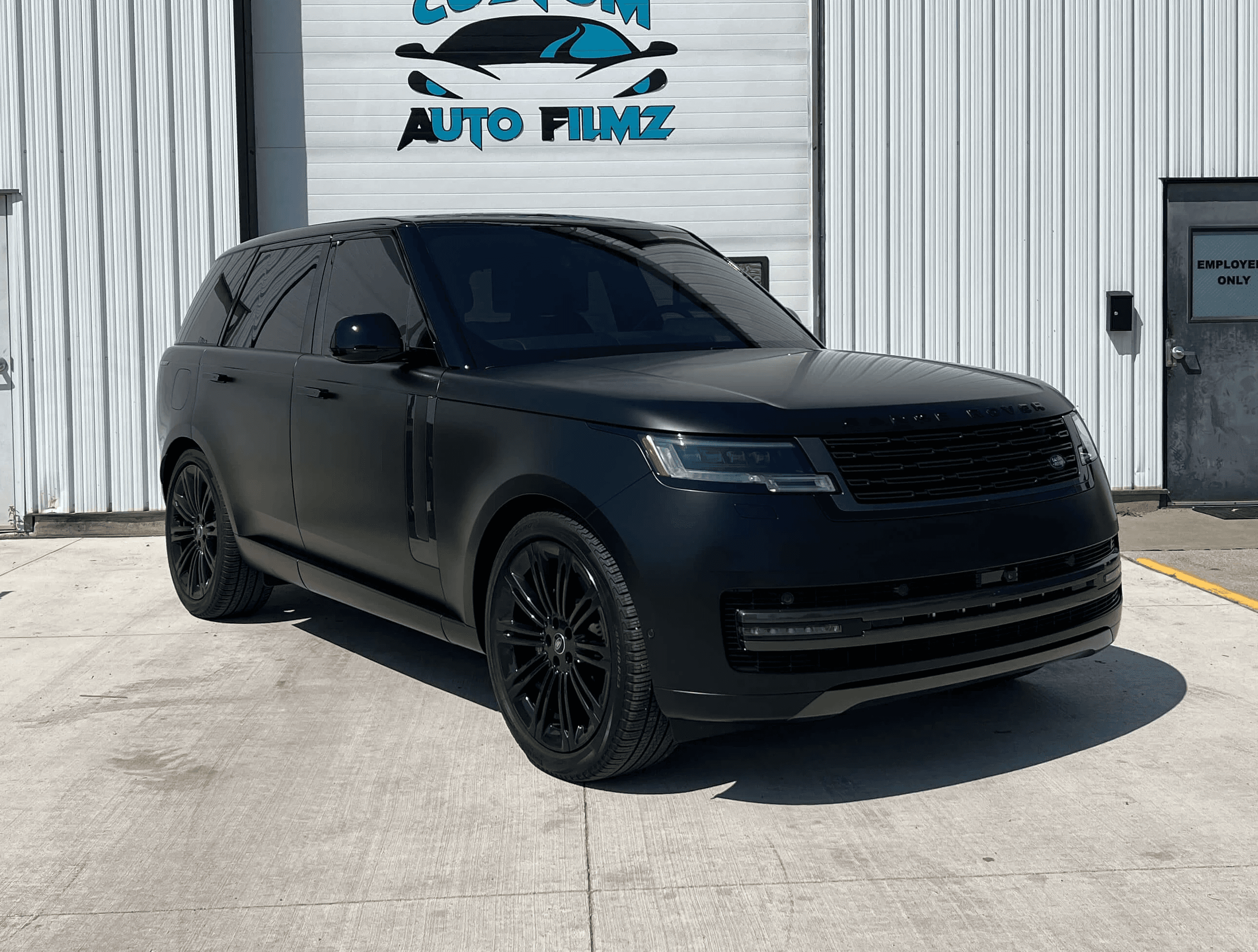 A matte black Range Rover is parked on a concrete driveway in front of a white industrial building.