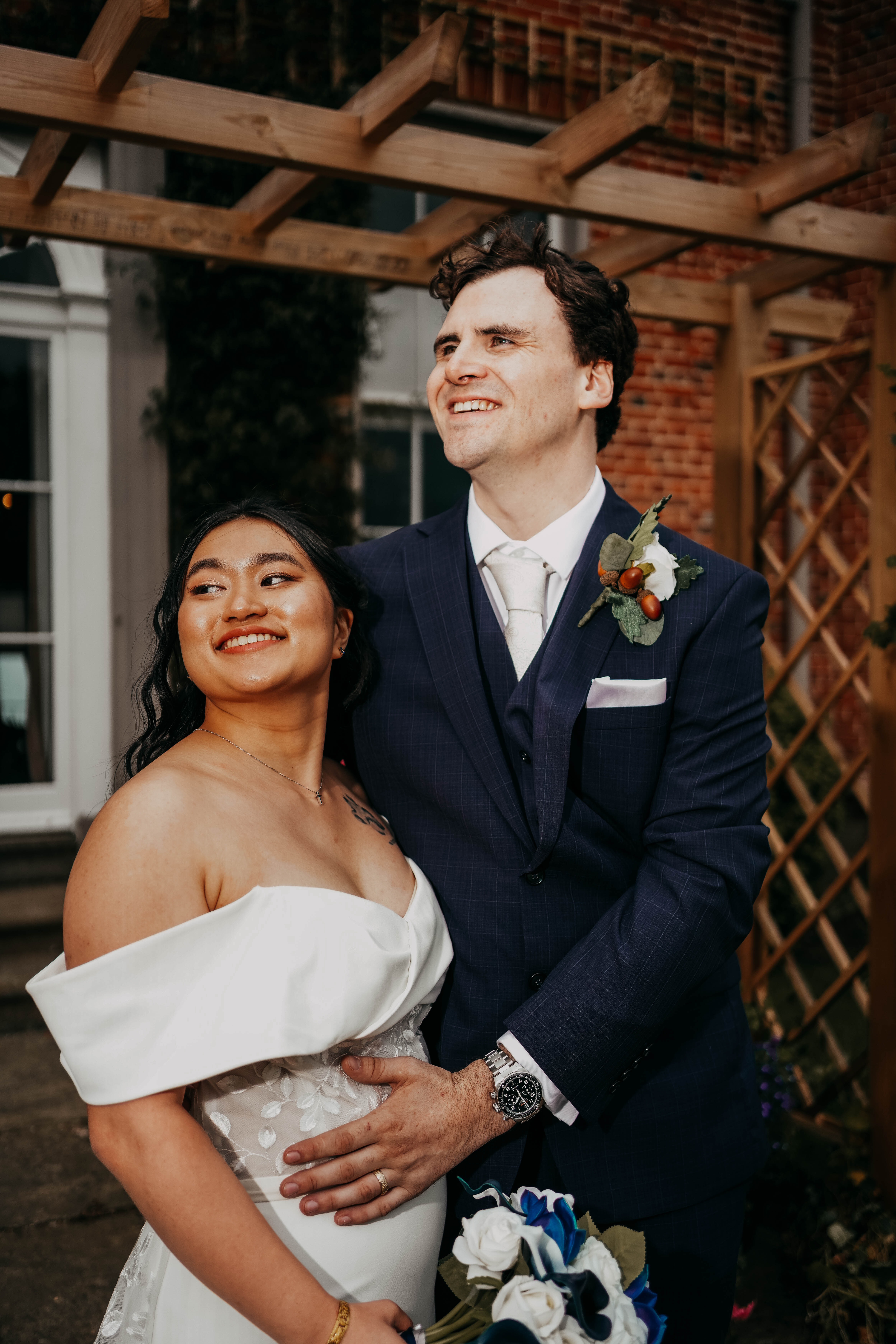The bride and groom pose together outdoors under a wooden pergola. The groom has his arm around the bride, and they are both smiling and looking away from the camera, their faces lit by warm sunlight.