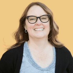 A smiling woman with shoulder-length blonde hair and black-rimmed glasses wearing a black cardigan over a light blue top with decorative neckline detailing, photographed against a warm yellow background.