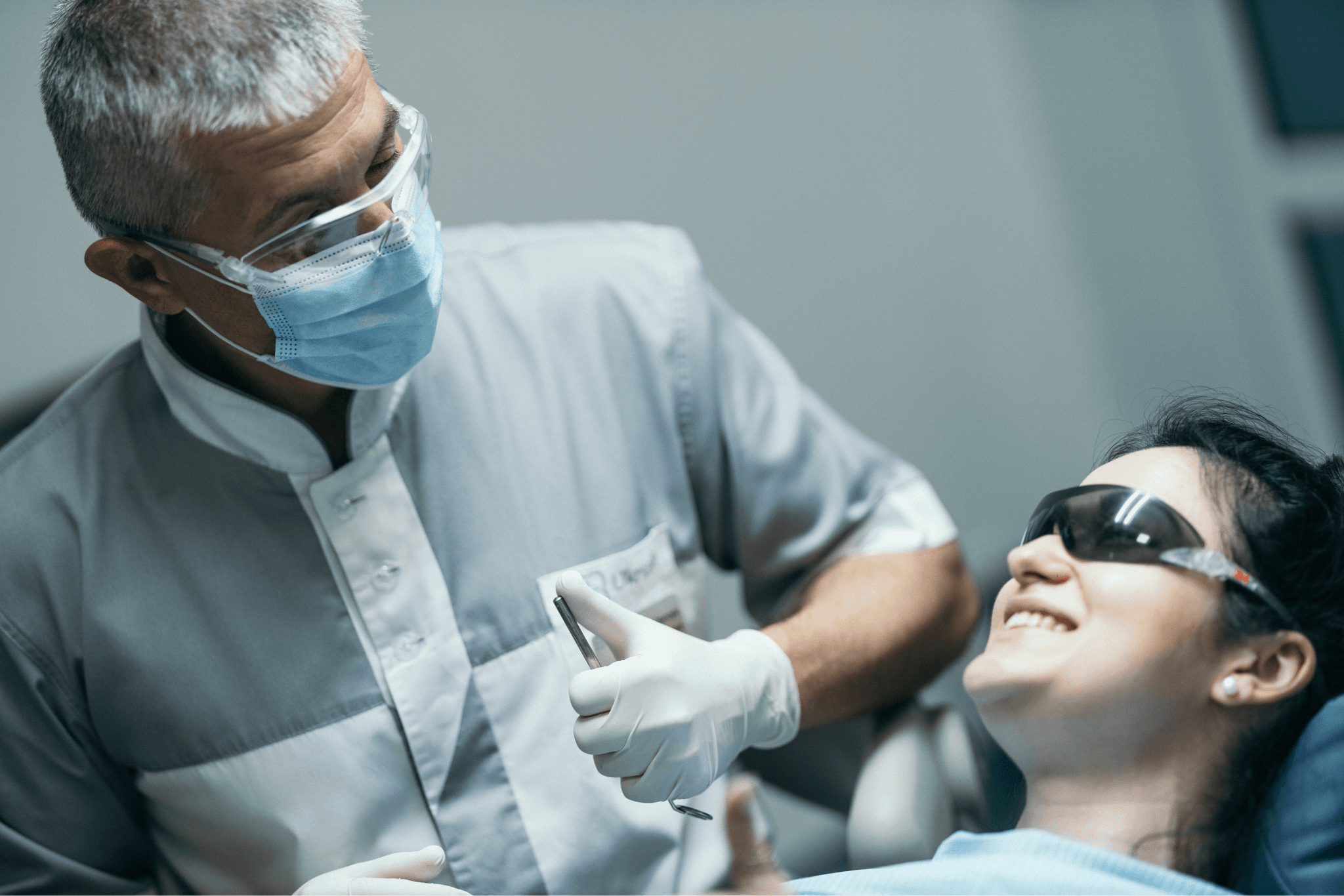 Стоматолог у захисній масці та окулярах схвалює результат лікування, консультує усміхнену пацієнтку, яка сидить у стоматологічному кріслі.