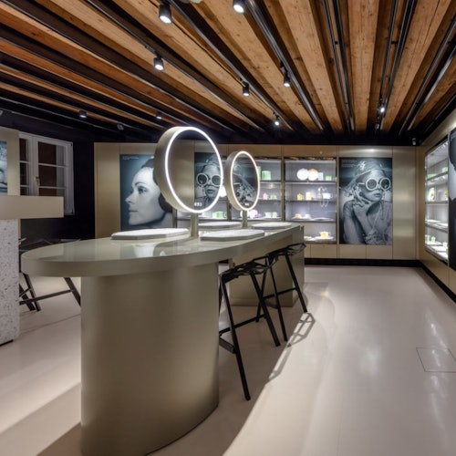 Showroom moderno com teto de madeira, espelhos iluminados em uma mesa central, prateleiras de exposição com itens e grandes retratos fotográficos.