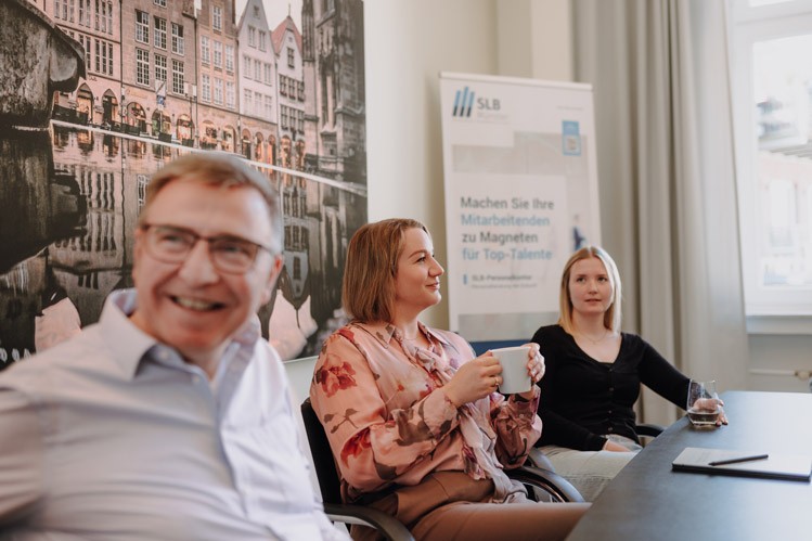 Besprechung bei SLB: Drei Personen sitzen an einem Tisch vor Präsentationswand und Flipchart.