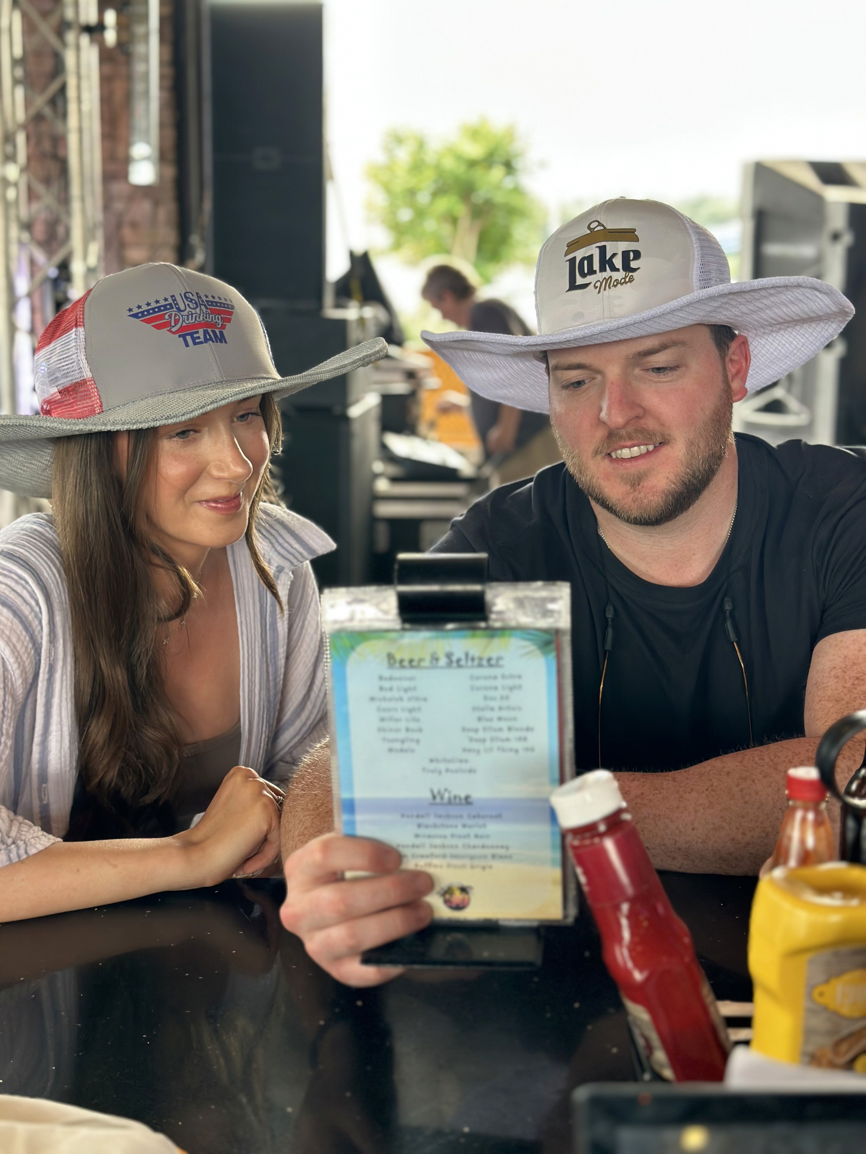 A man and woman wearing wide-brimmed hats sit at an outdoor bar table reading a menu, with ketchup and mustard bottles visible in the foreground.