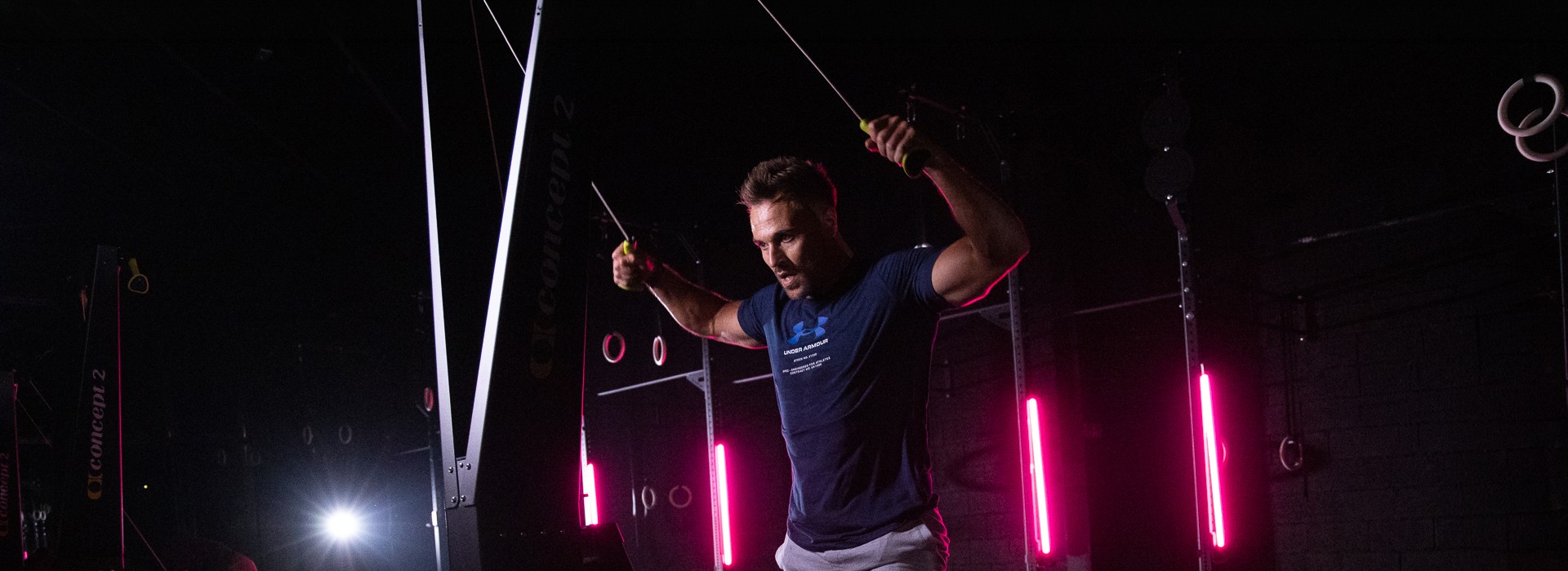 A male athlete pulling down on a Concept2 SkiErg in a dark gym, lit by magenta strip lights.