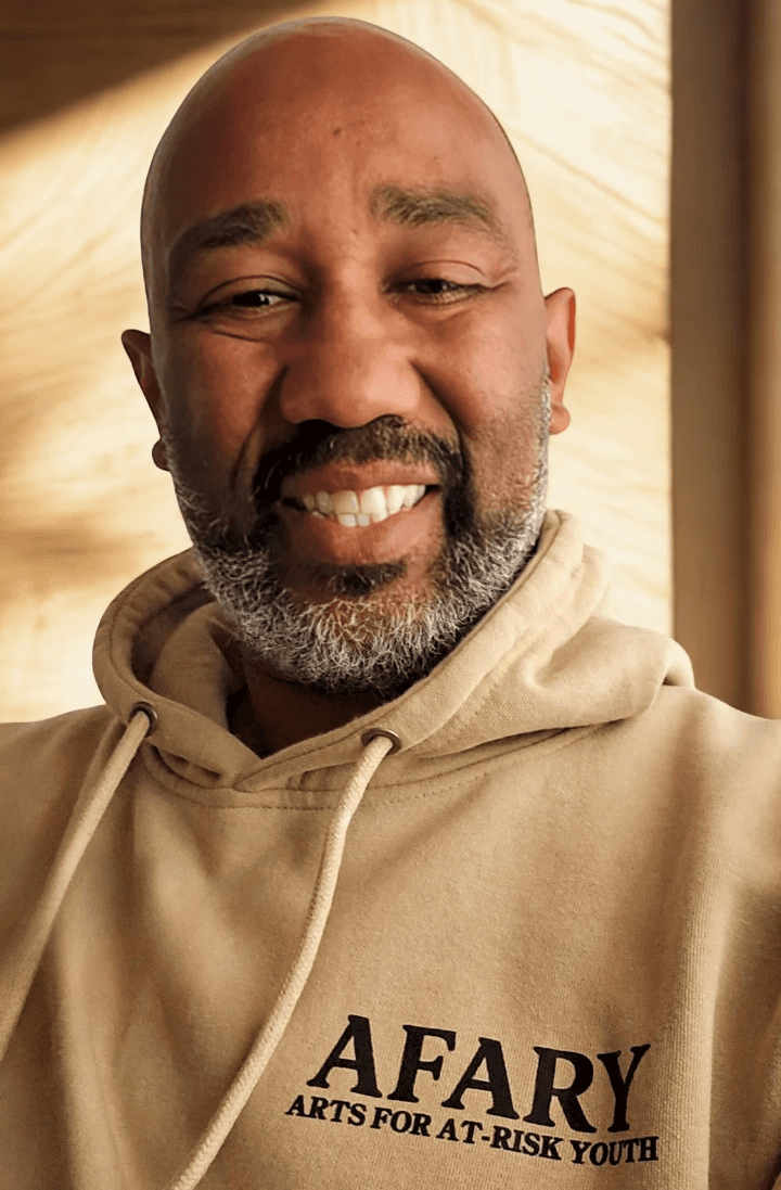 Smiling woman in beige yoga outfit sitting in natural light with green plants in the background.