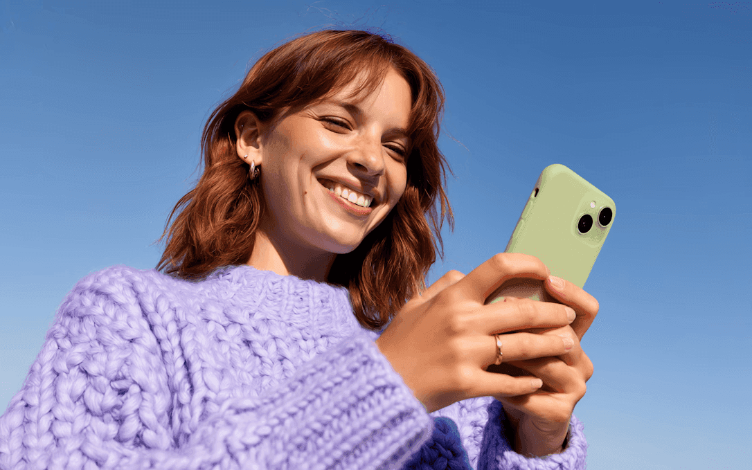 A woman wearing a purple sweater holds a green smartphone and smiles warmly under a clear blue sky, conveying happiness and connectivity.