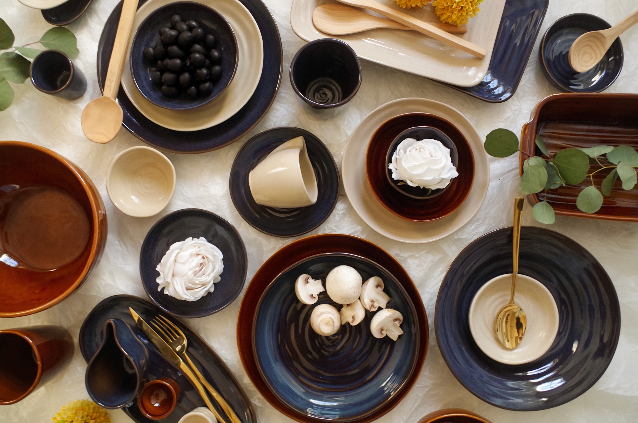 Vue du dessus d'une collection de céramiques bleu nuit et brun réactif, assiettes, bols, mugs et plats assortis avec champignons, olives et fleurs blanches