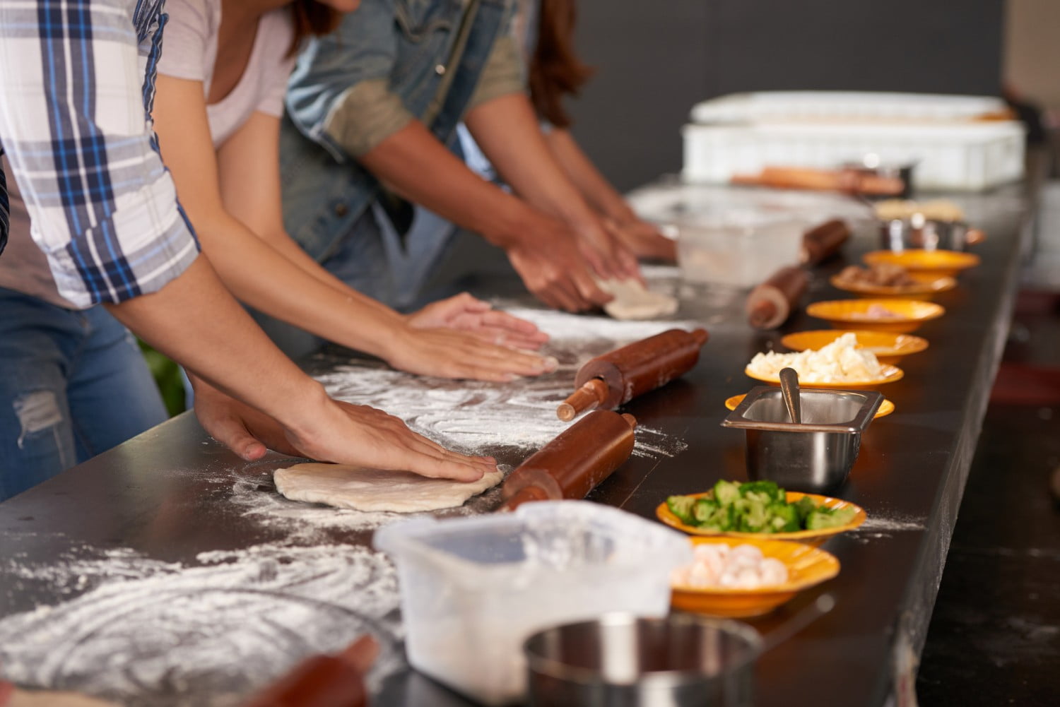 A person kneads fresh dough on a wooden kitchen counter dusted with flour, with pottery and a plant visible in the background, evoking a rustic baking atmosphere.