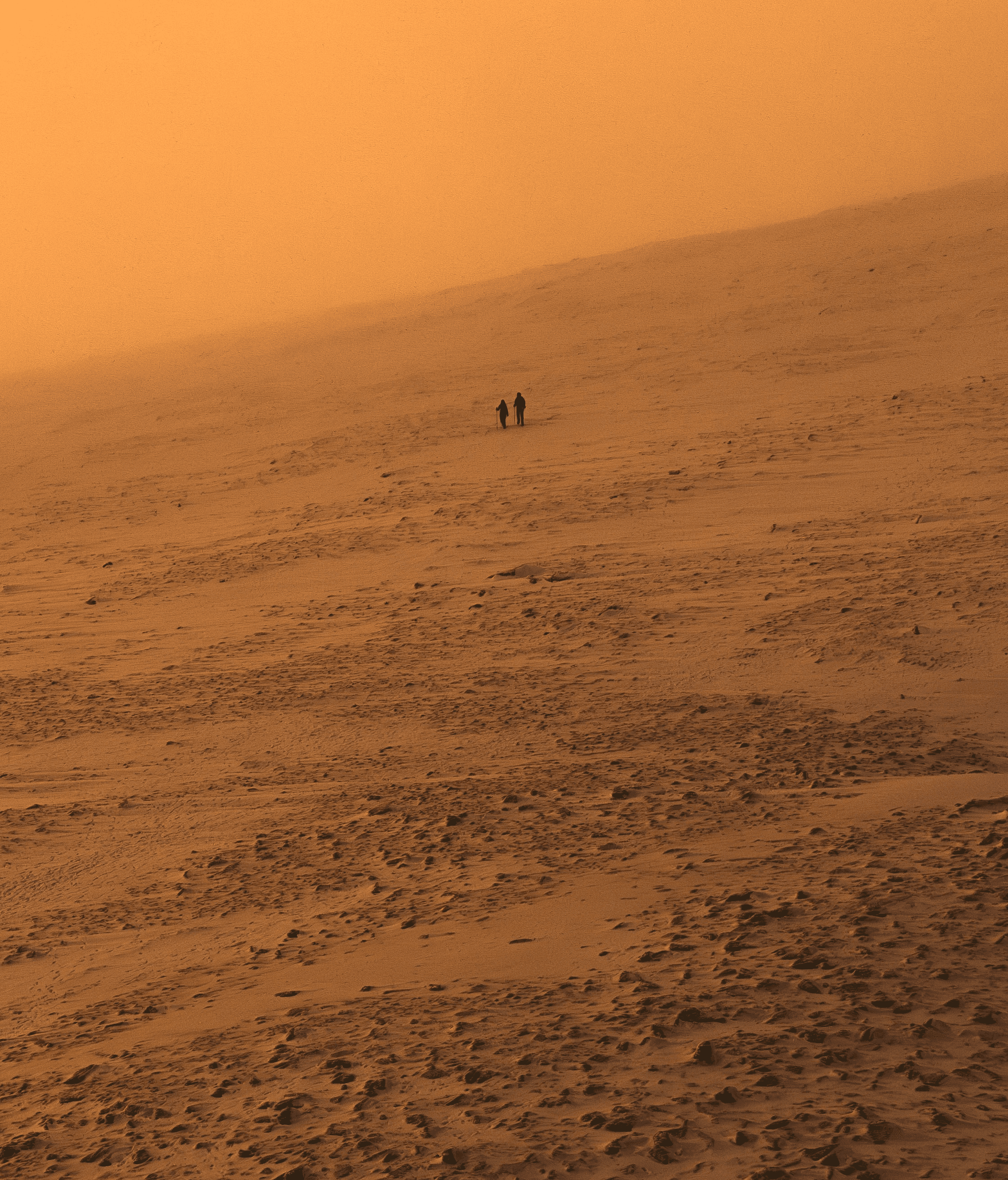 A long-distance image of two people dressed in black walking through a desert sand storm.