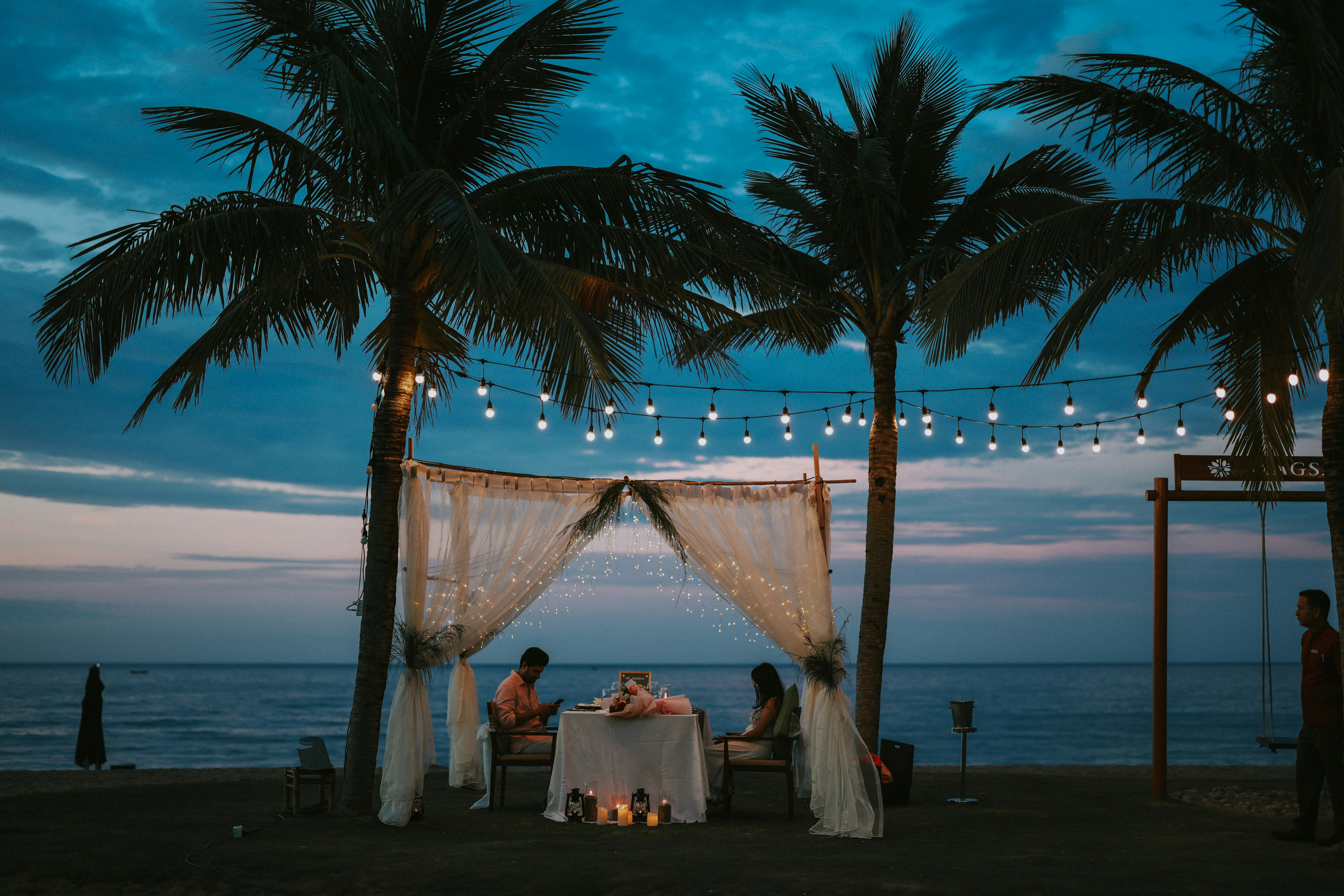 Couple dining under palm trees at dusk with string lights.