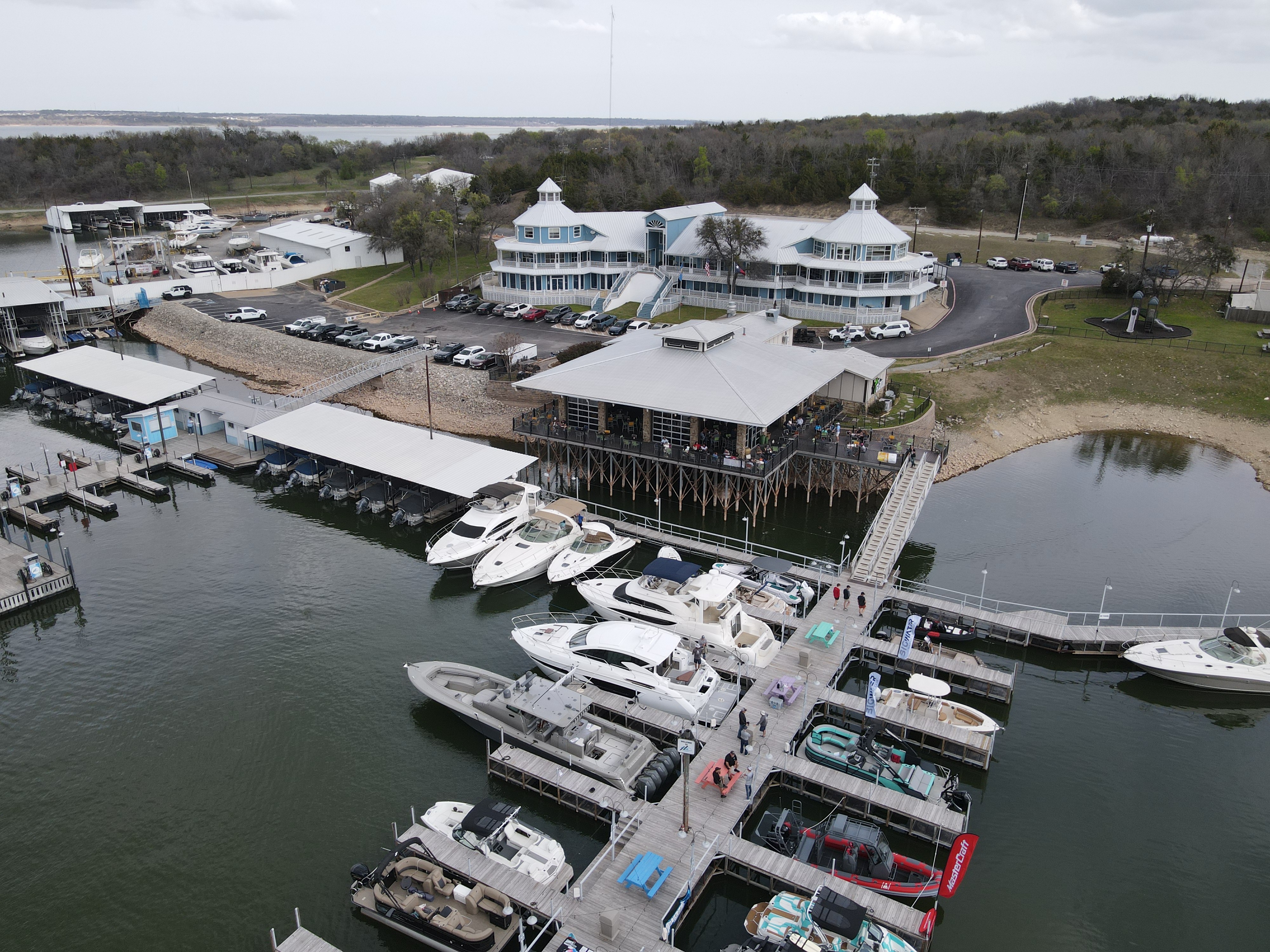 Aerial view of a bustling marina featuring several boats docked at wooden piers, with a large, elevated waterfront building and a modern white structure surrounded by parked cars in the background.